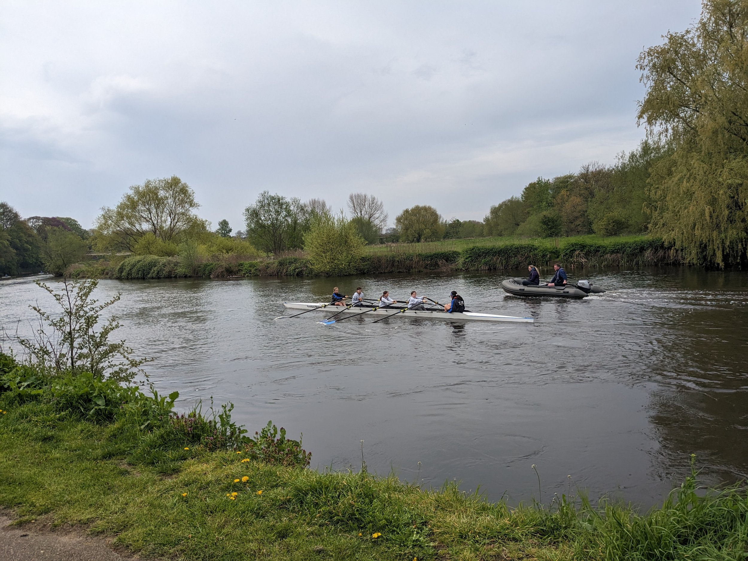 A group of seven people rowing a boat on a river while a small motorized boat with two people follows nearby. The river is surrounded by trees and greenery, with an overcast sky overhead.