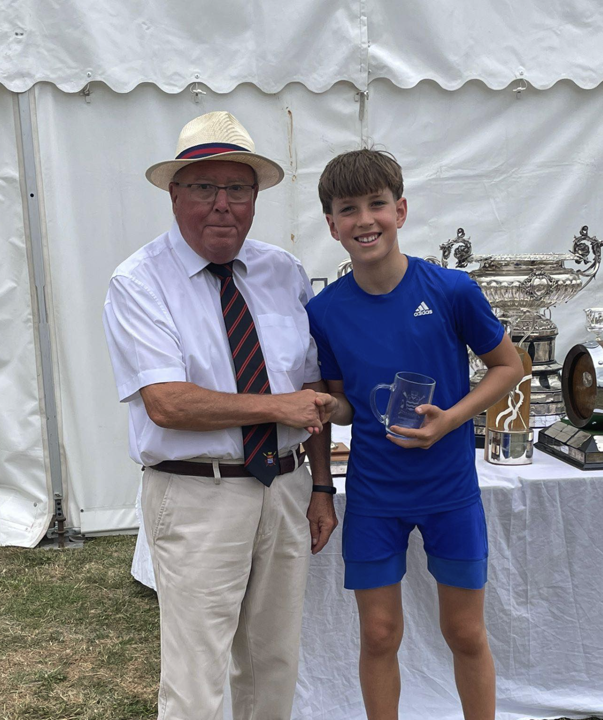 An older man wearing a straw hat, glasses, a white shirt, and a striped tie shakes hands with a young boy in a blue sports shirt, holding a glass trophy, in front of a table with silver trophies inside a white tent.