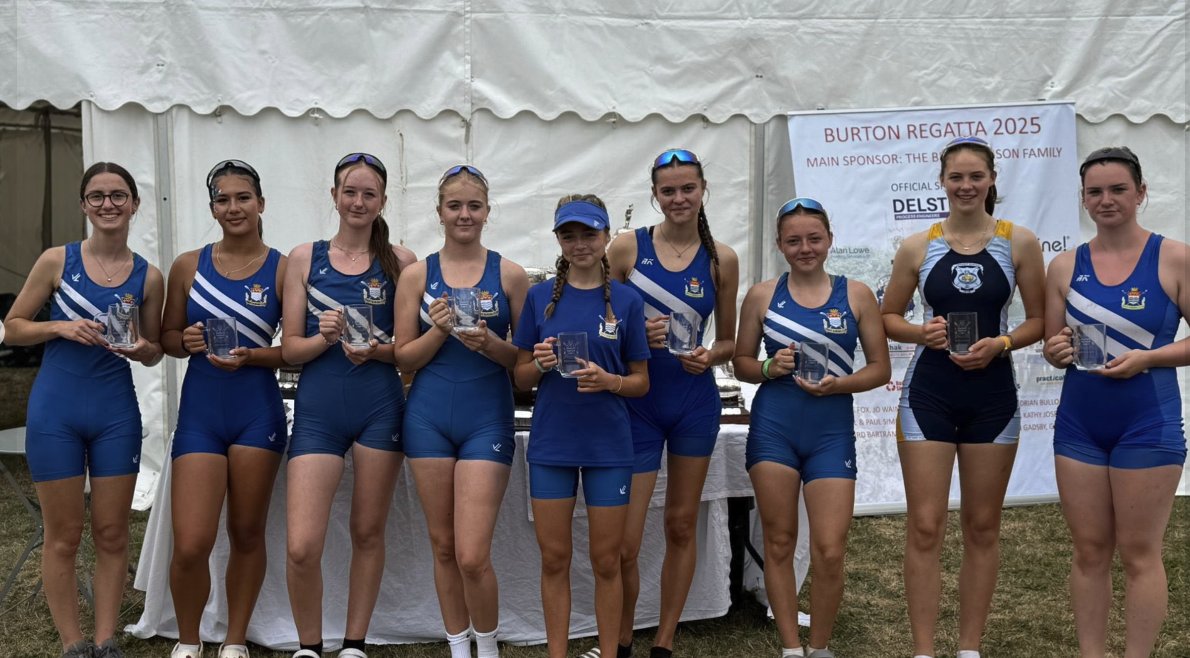 Group of young women in blue athletic uniforms holding clear glass awards at the Burton Regatta 2025 event, standing in front of a white tent with a banner.