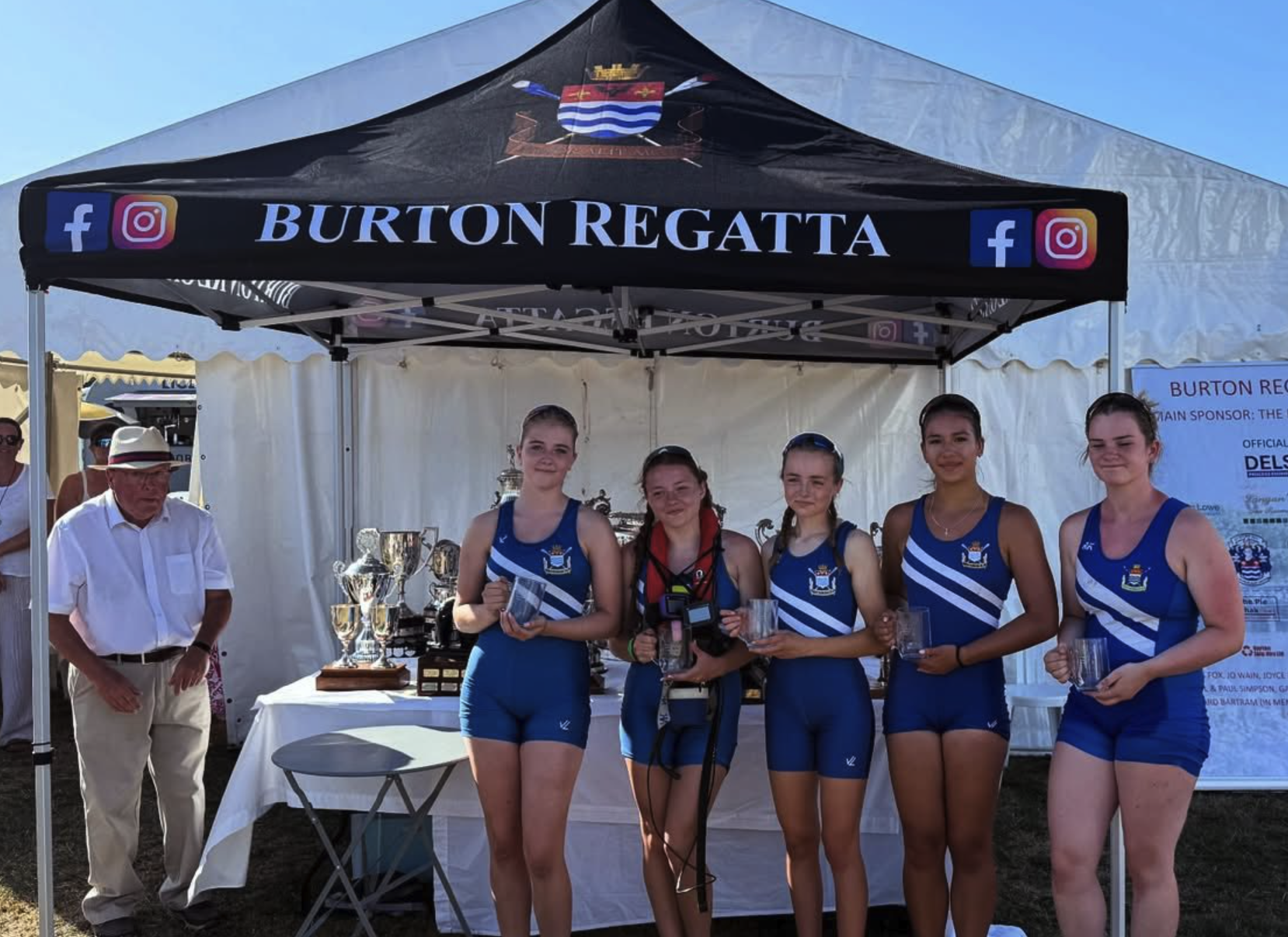 Six young women in blue athletic uniforms posing with transparent mugs under a black canopy labeled 'Burton Regatta' with social media icons. An older man in a white shirt and hat stands to the left, with trophies and awards on a table behind them.