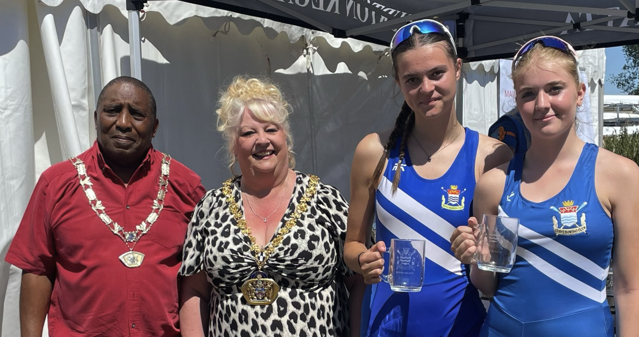 Four people standing outdoors, two men on the left wearing necklaces, possibly ceremonial. The two women on the right are in blue athletic uniforms with a crest, holding glass mugs. They are under a tent, with a marina and boats visible in the backgr