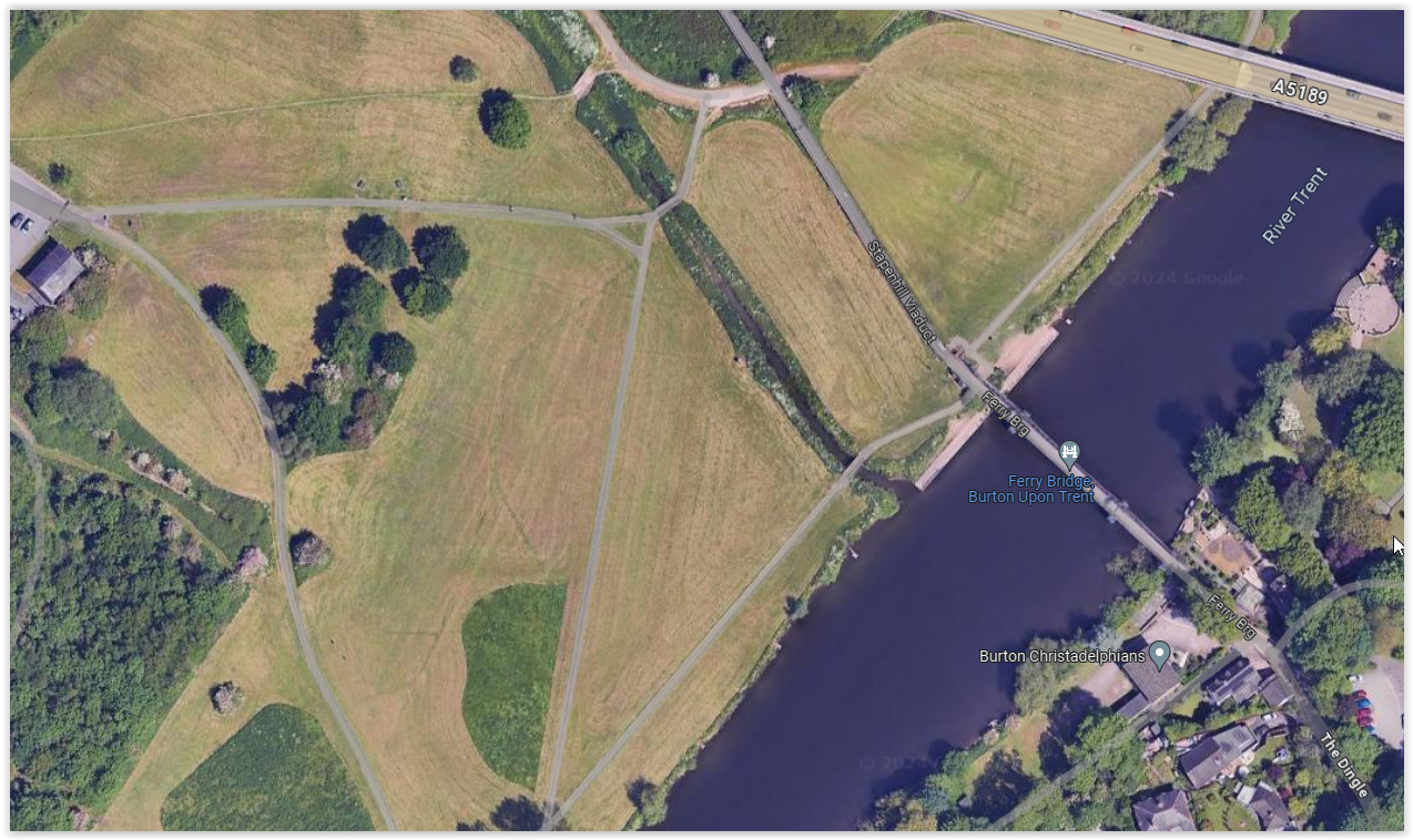 Aerial view of a park with walking paths, green grass, trees, and a river with a bridge crossing over it.