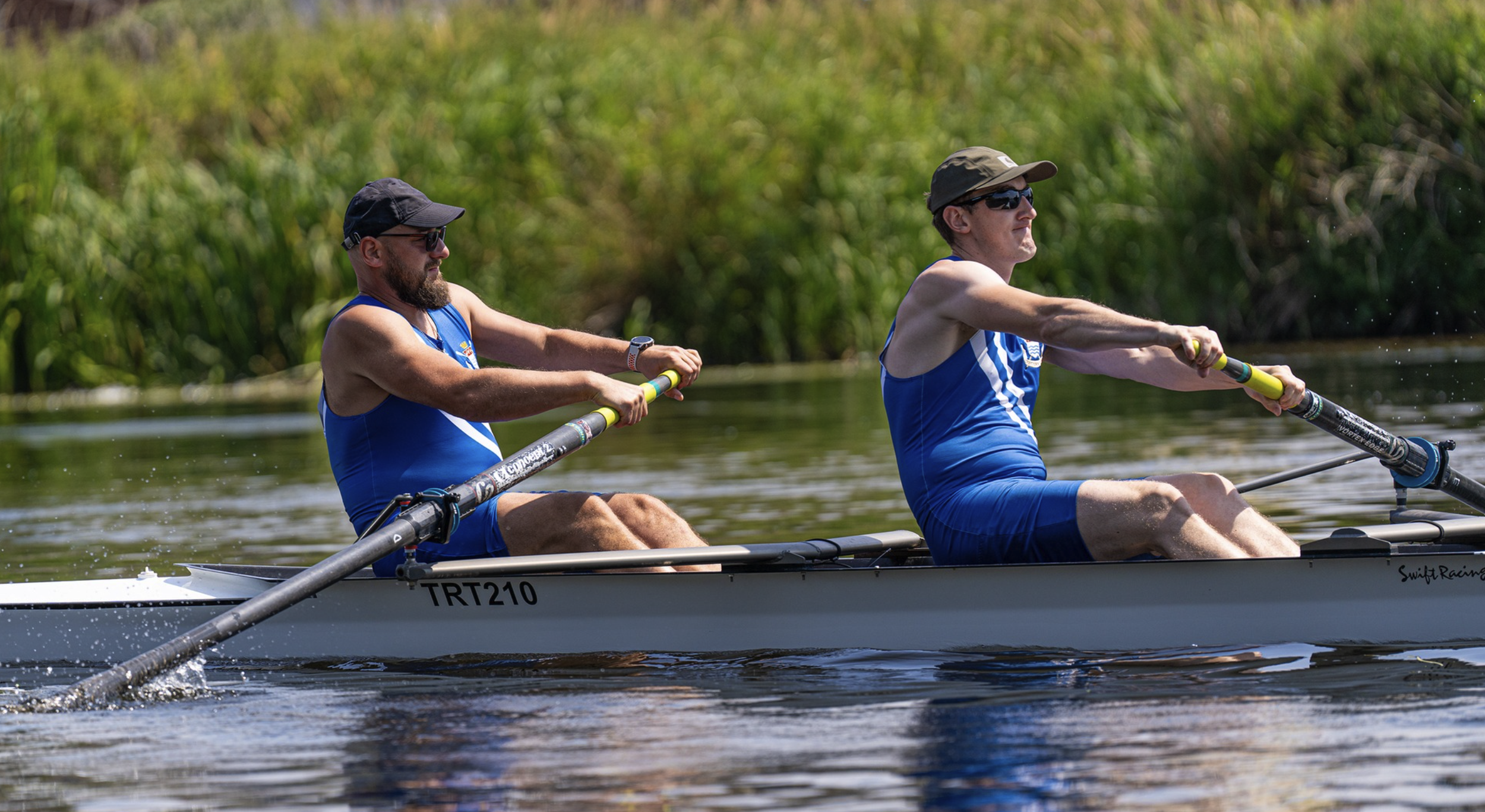Two men from Trent Rowing Club rowing a boat on the water, both wearing blue athletic gear, hats, and sunglasses.