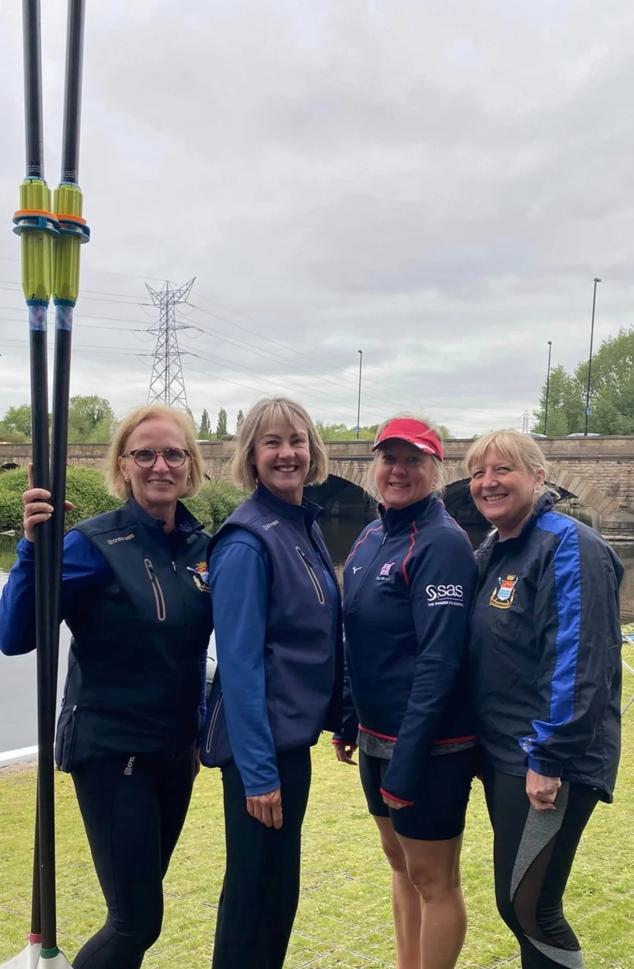 Four women in sports uniforms standing outdoors near a bridge, holding paddles, smiling at the camera.