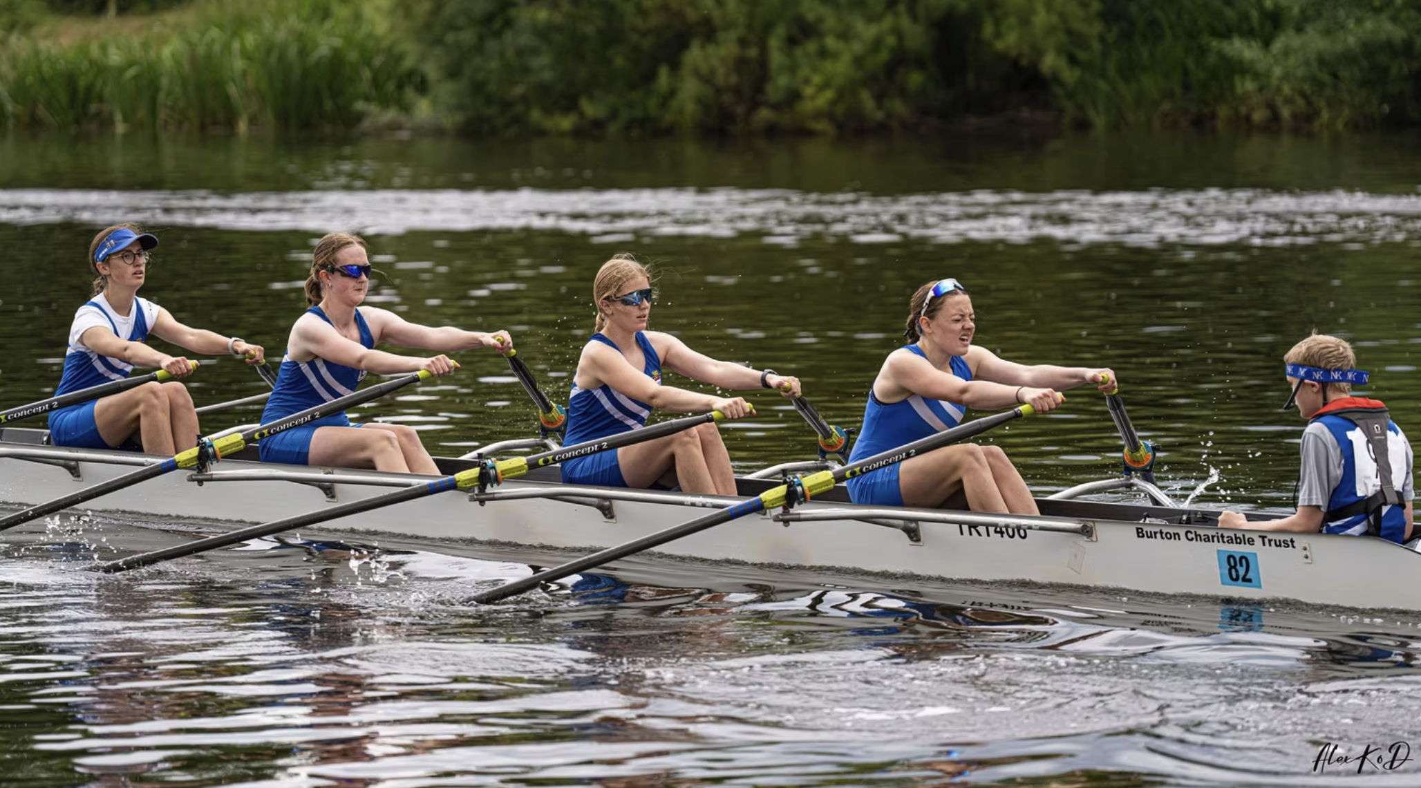 Four women in blue and white uniforms rowing a boat on a lake, with a person at the front steering, while a person at the back supervises, on a cloudy day.