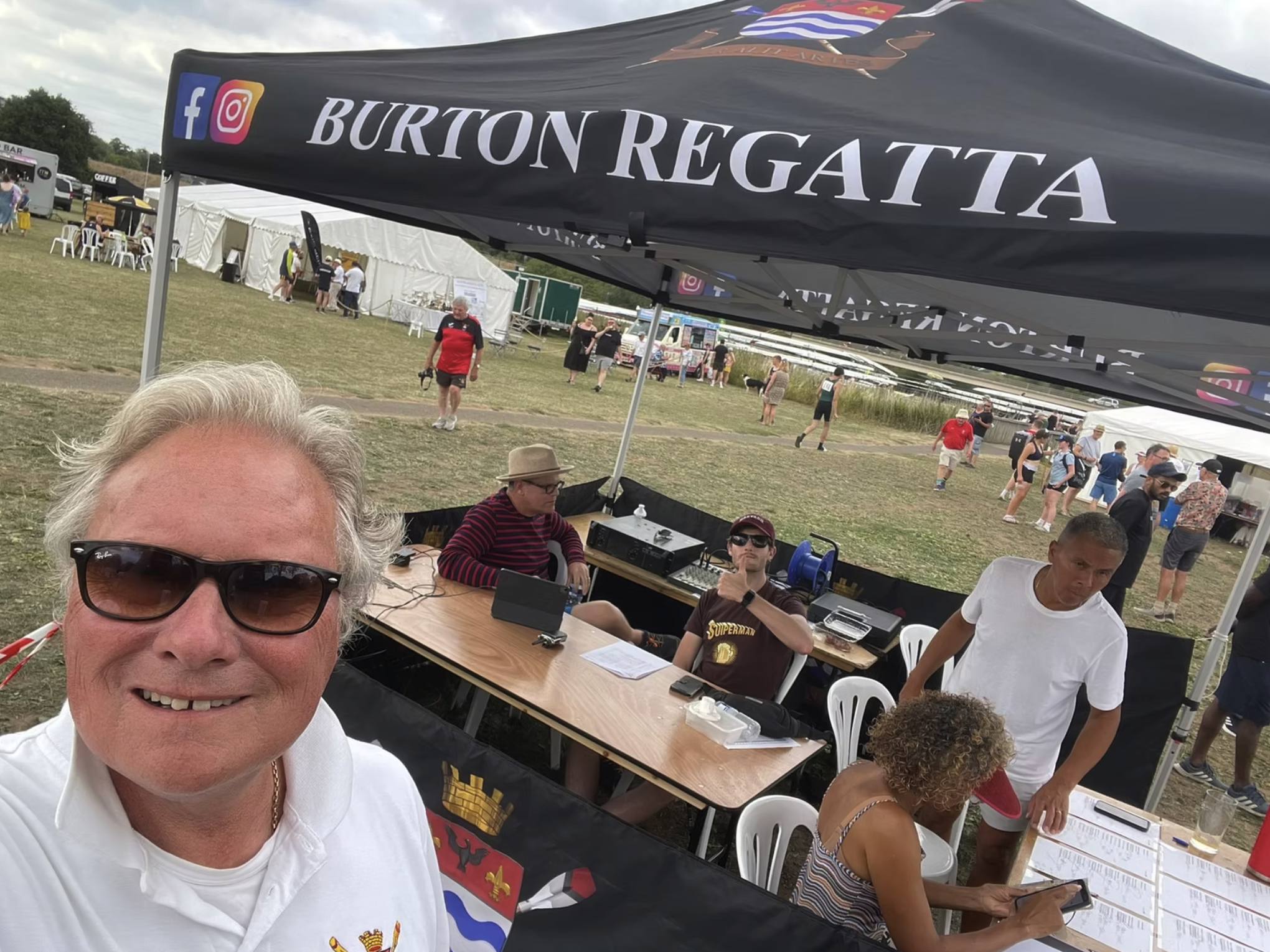 A man with white hair and sunglasses taking a selfie at an outdoor event, with a large black canopy displaying the name 'Burton Regatta' and social media icons. There are people sitting at tables and walking in the background.