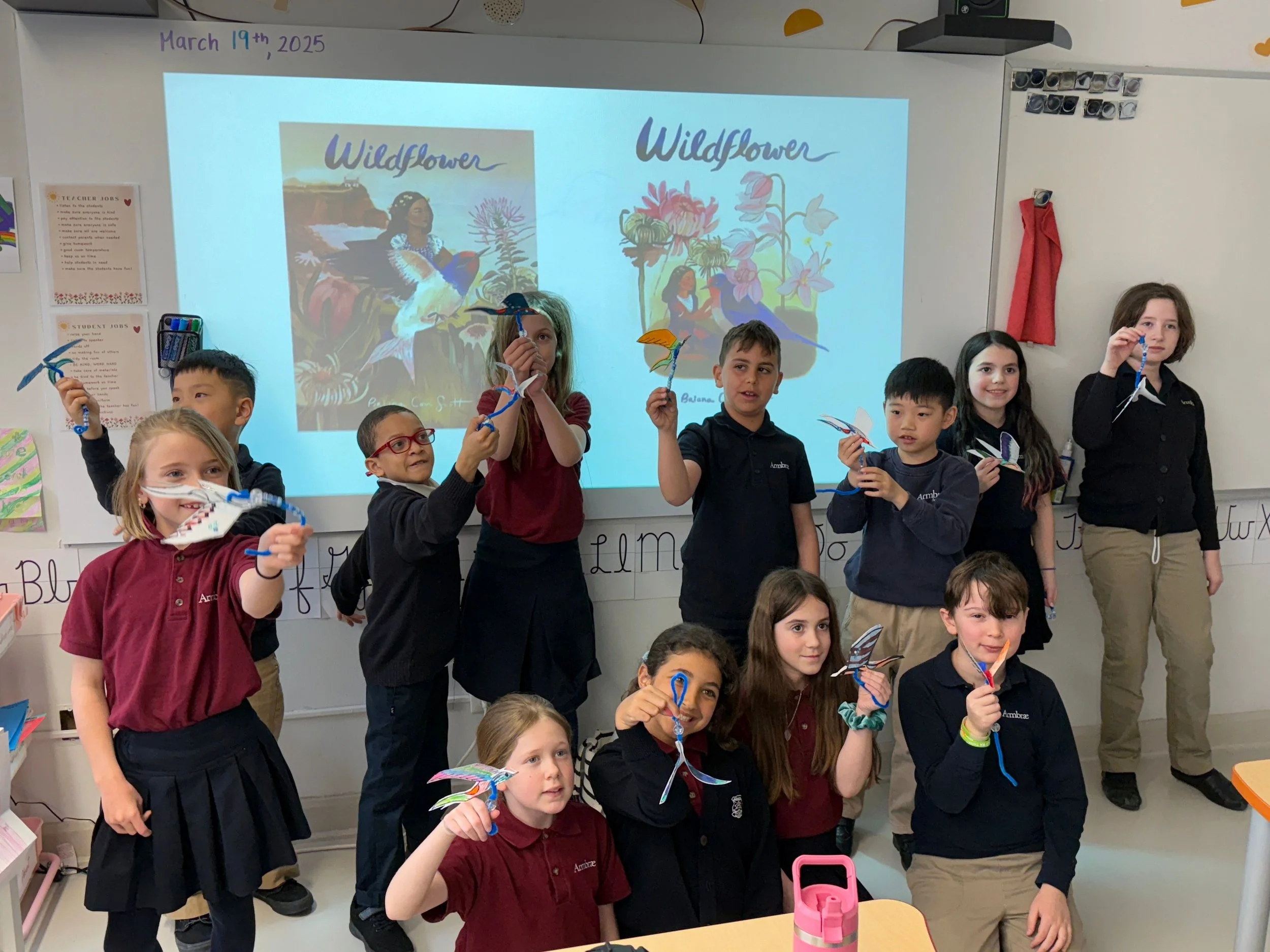 A group of children in a classroom holding paper windmill crafts, standing in front of a whiteboard with a projected image and text. The children are smiling and appear to have completed a craft activity related to a book or story titled 'Wildflower.'