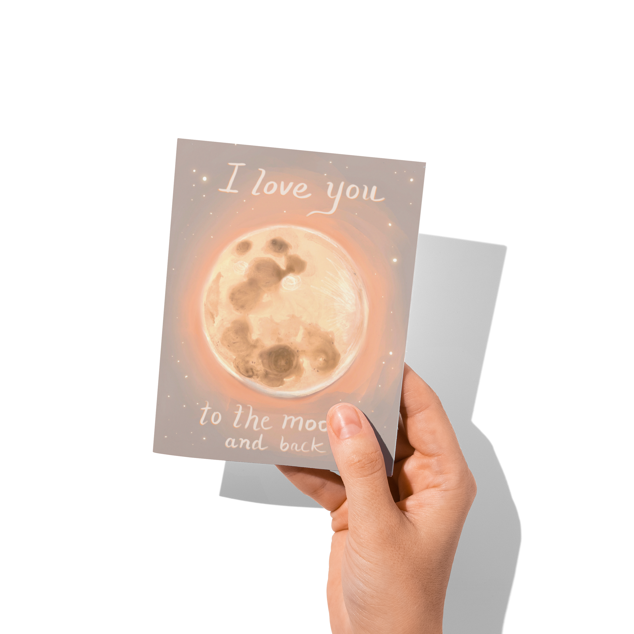 Hand holding a greeting card illustrated with a glowing full moon and the phrase “I love you to the moon and back” – artwork by Briana Corr Scott