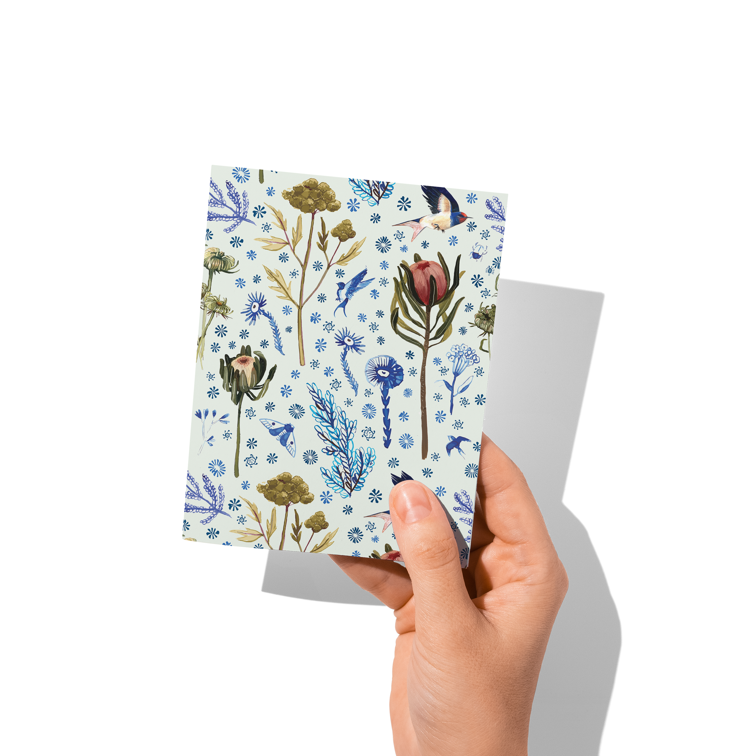 Hand holding a greeting card featuring a soft botanical pattern of protea, seaweed, feathers, and wildflowers in blue and green tones – by Briana Corr Scott