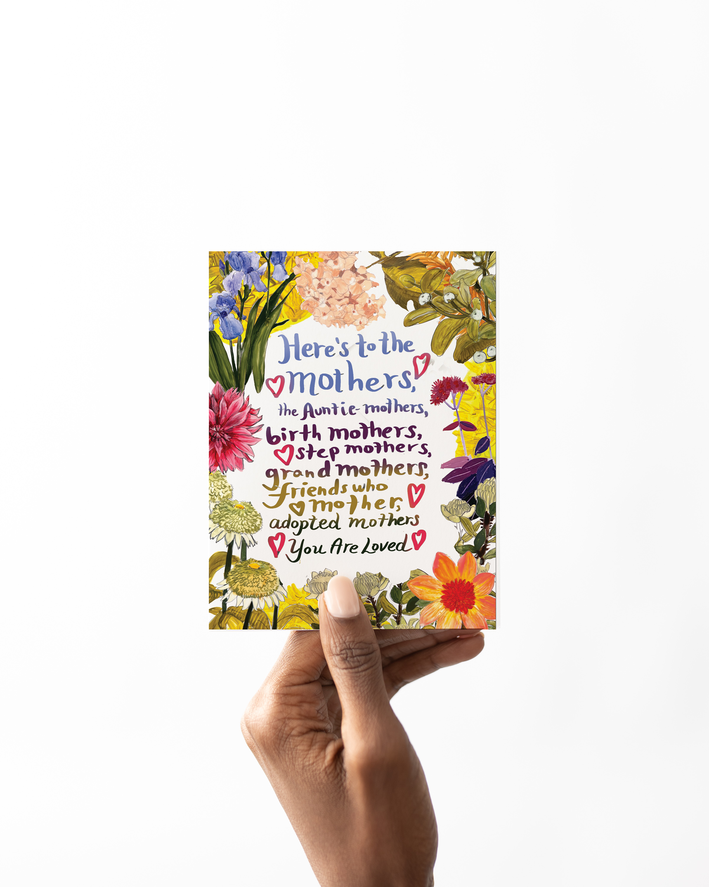 Hand holding a greeting card that reads “Here’s to the mothers...” surrounded by colorful flowers – celebrates all kinds of moms, illustrated by Briana Corr Scott