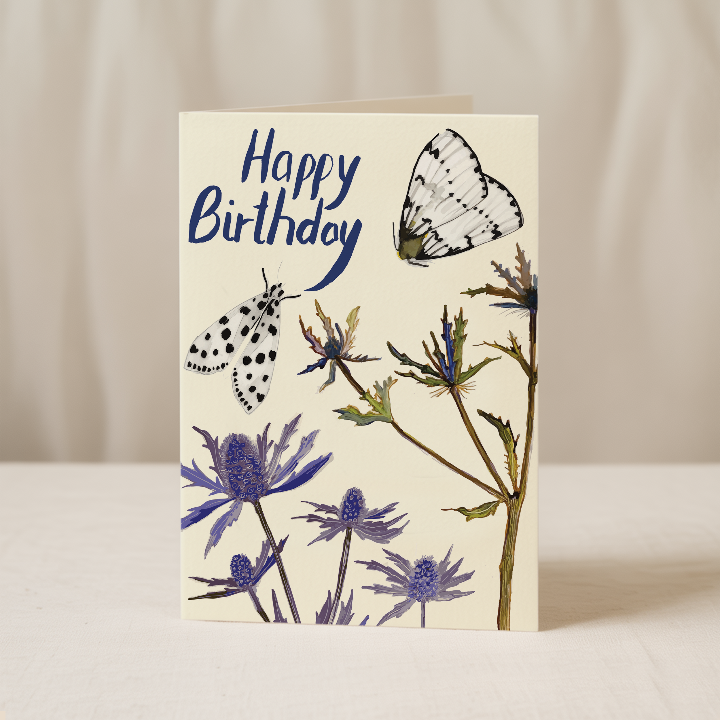 Greeting card with “Happy Birthday” hand-lettered in blue at the top, featuring two black-and-white moths and purple thistle-like flowers against a cream background.