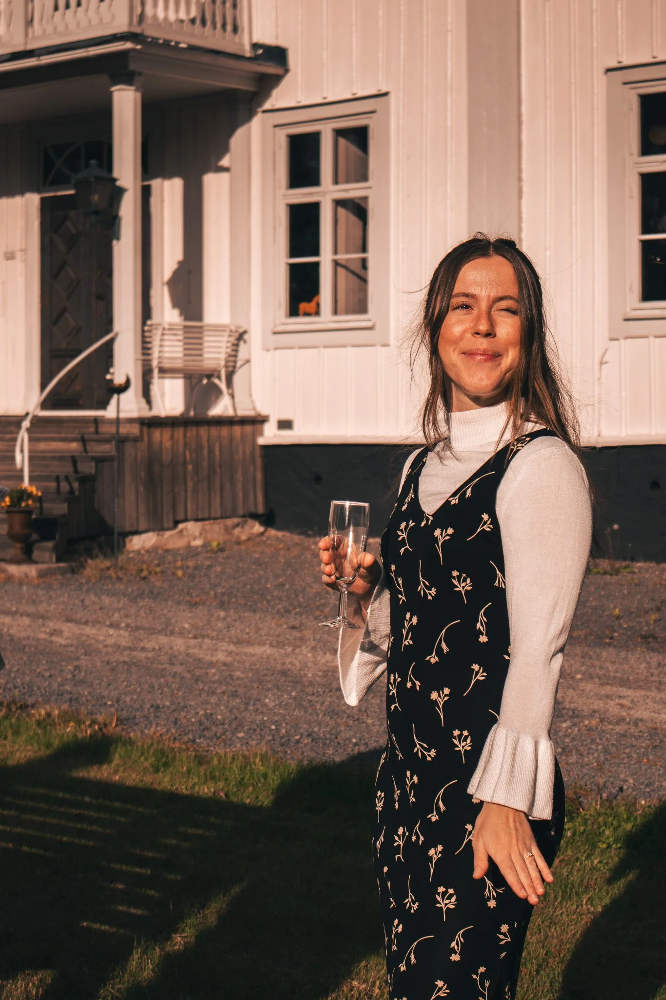 A woman smiling and holding a champagne flute outside a white wooden house during sunset.