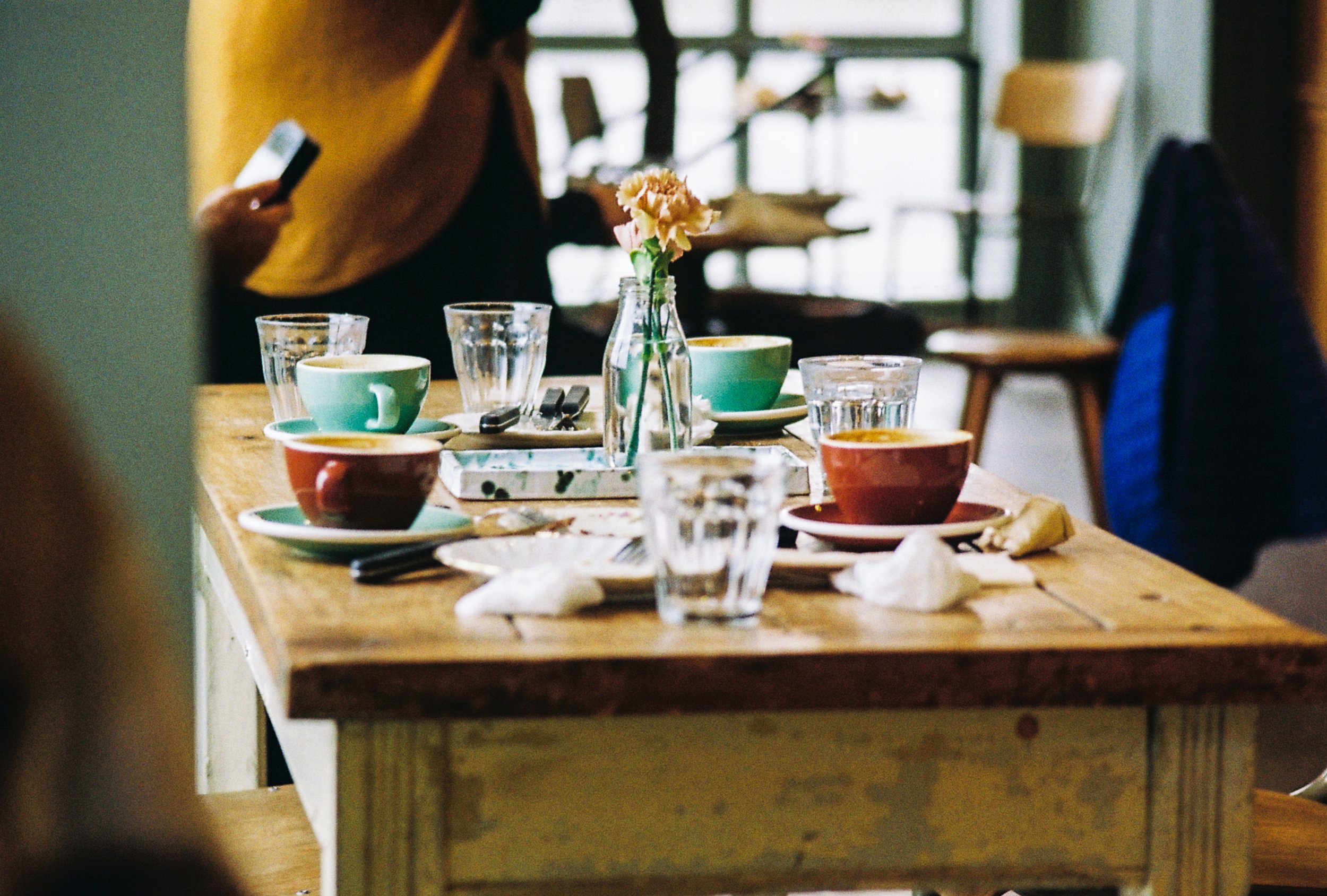A wooden table set with colorful cups of coffee, glasses of water, napkins, and a vase with pink flowers in a cozy cafe.