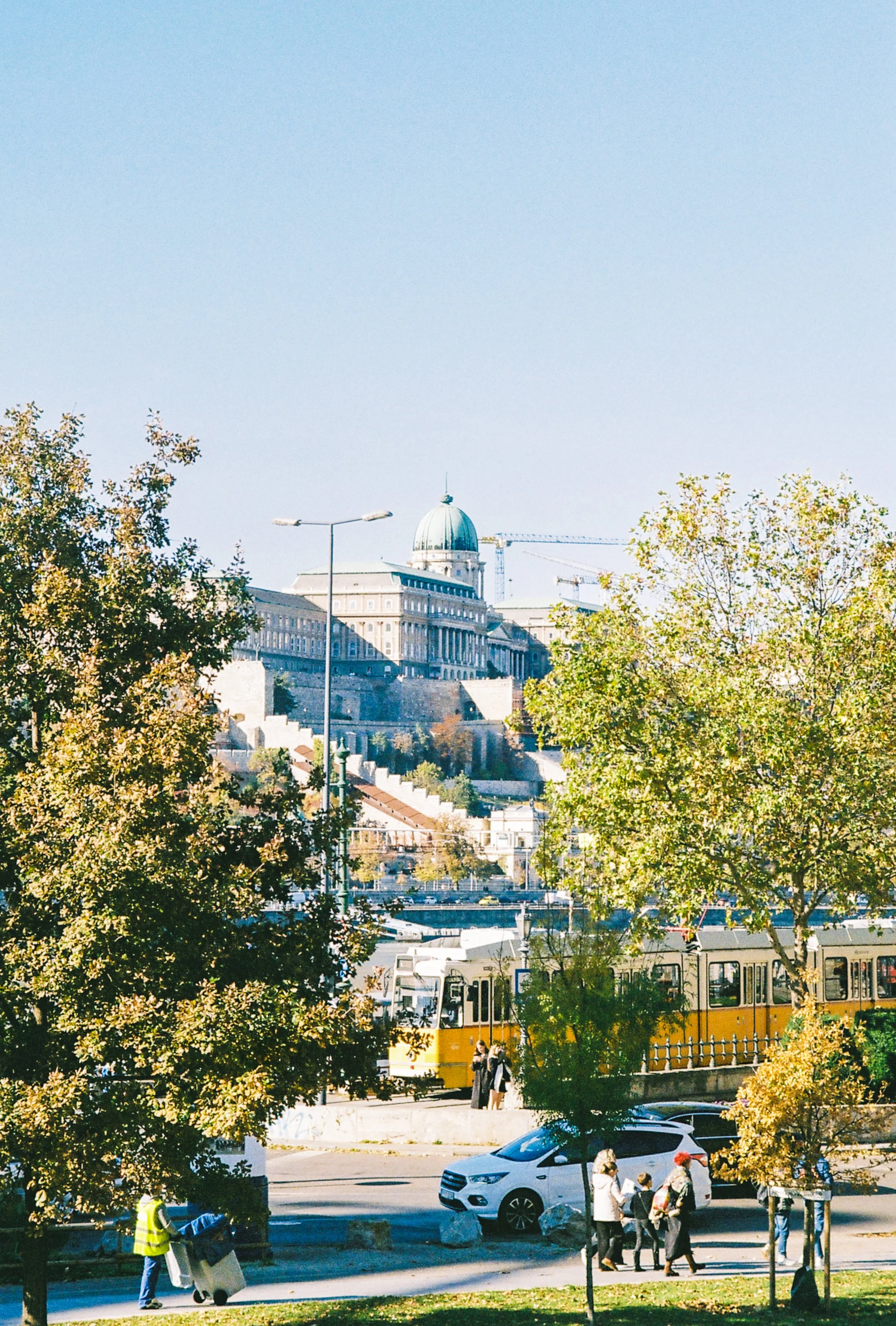 Cityscape with a historic building on a hill, a train, trees, cars, and pedestrians.