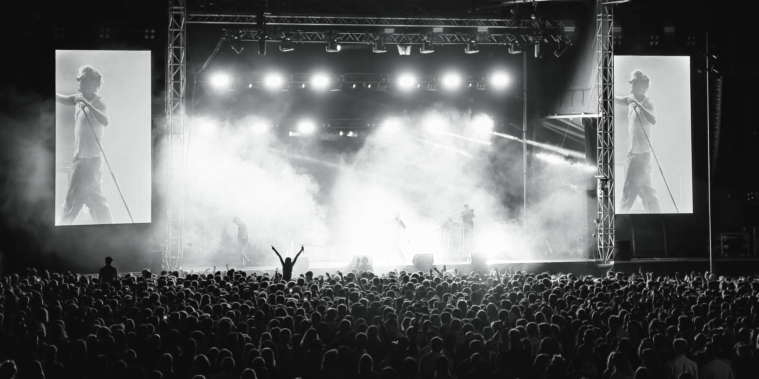 Black and white photo of a concert with a large crowd, stage lights, and a performer on stage with two large screens on each side showing close-ups of the performer.