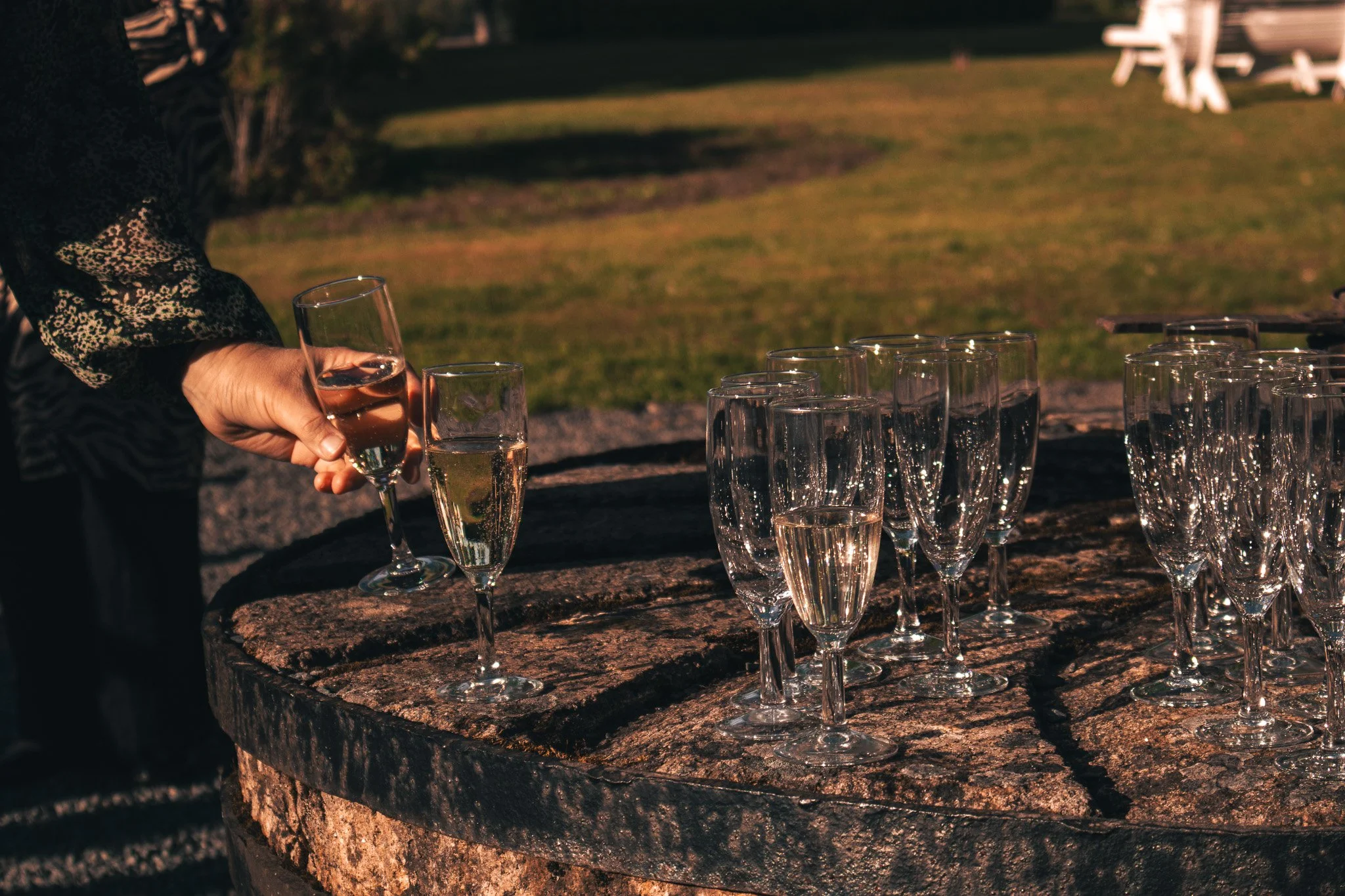 Person pouring champagne into a flute on a stone barrel with multiple filled glasses in an outdoor setting.
