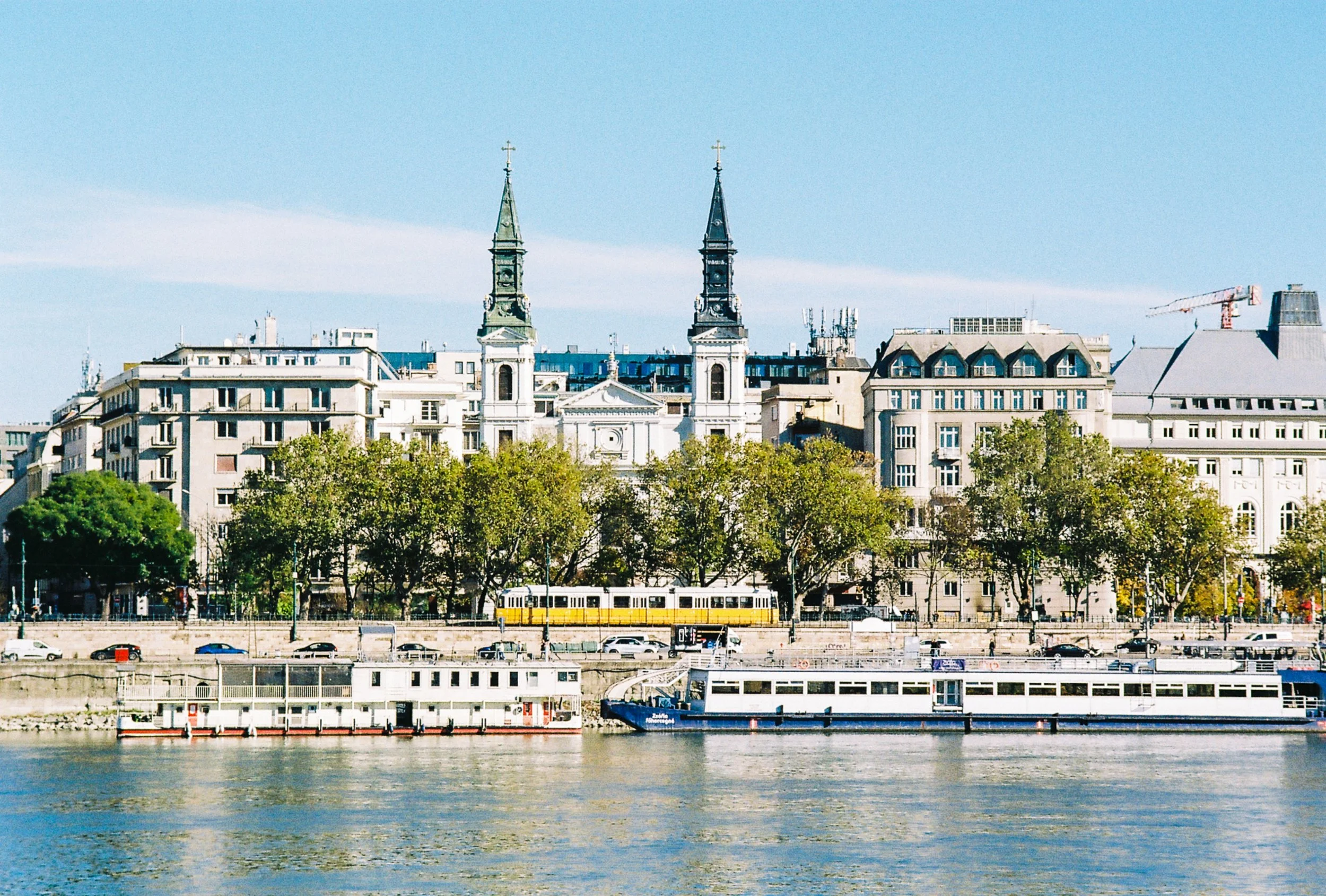 A cityscape view of buildings across a river, featuring two church steeples in the center, with trees, boats on the river, and a clear blue sky.
