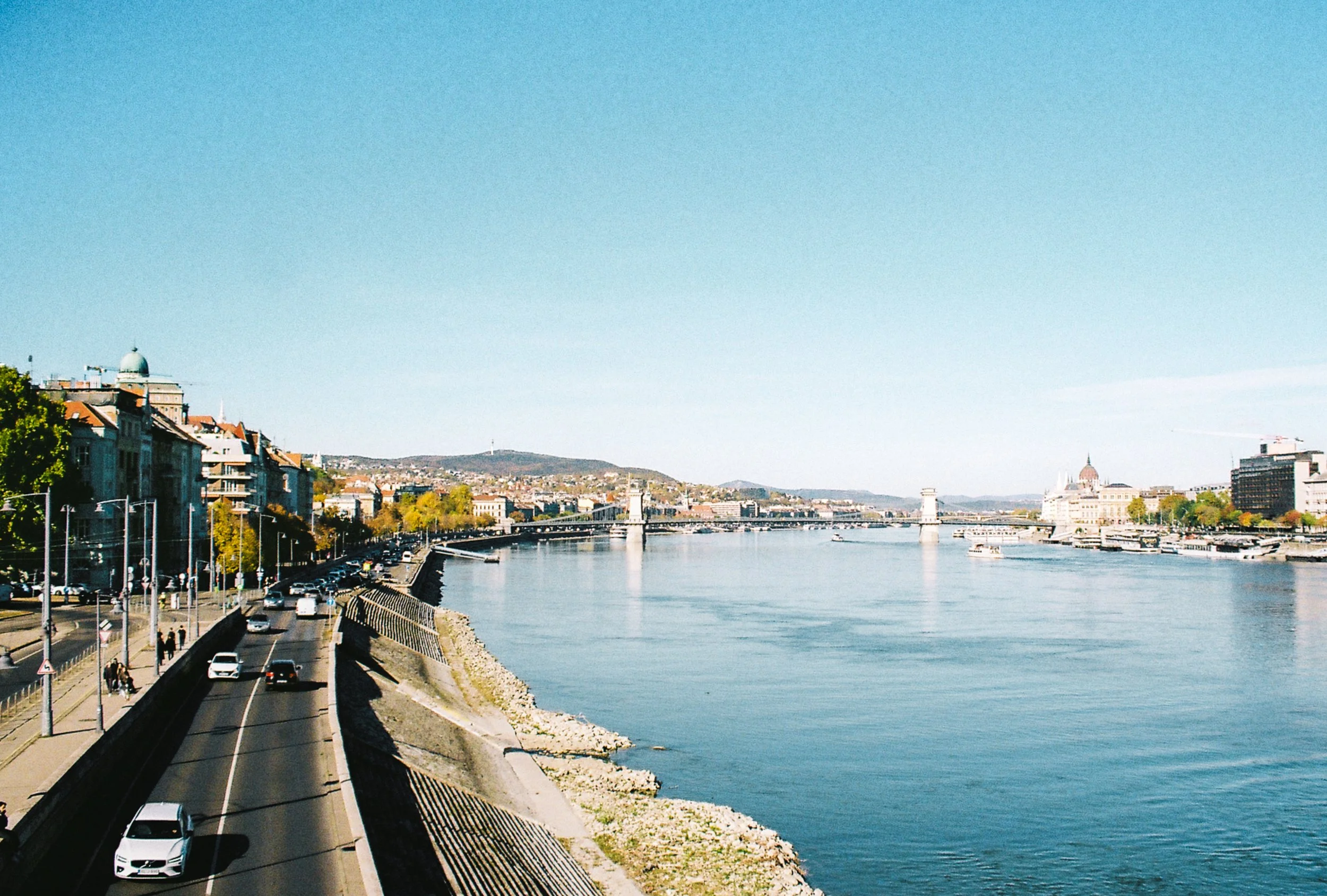 River with boats and cityscape on both sides, blue sky, distant hills, and structures including bridges and historic buildings.