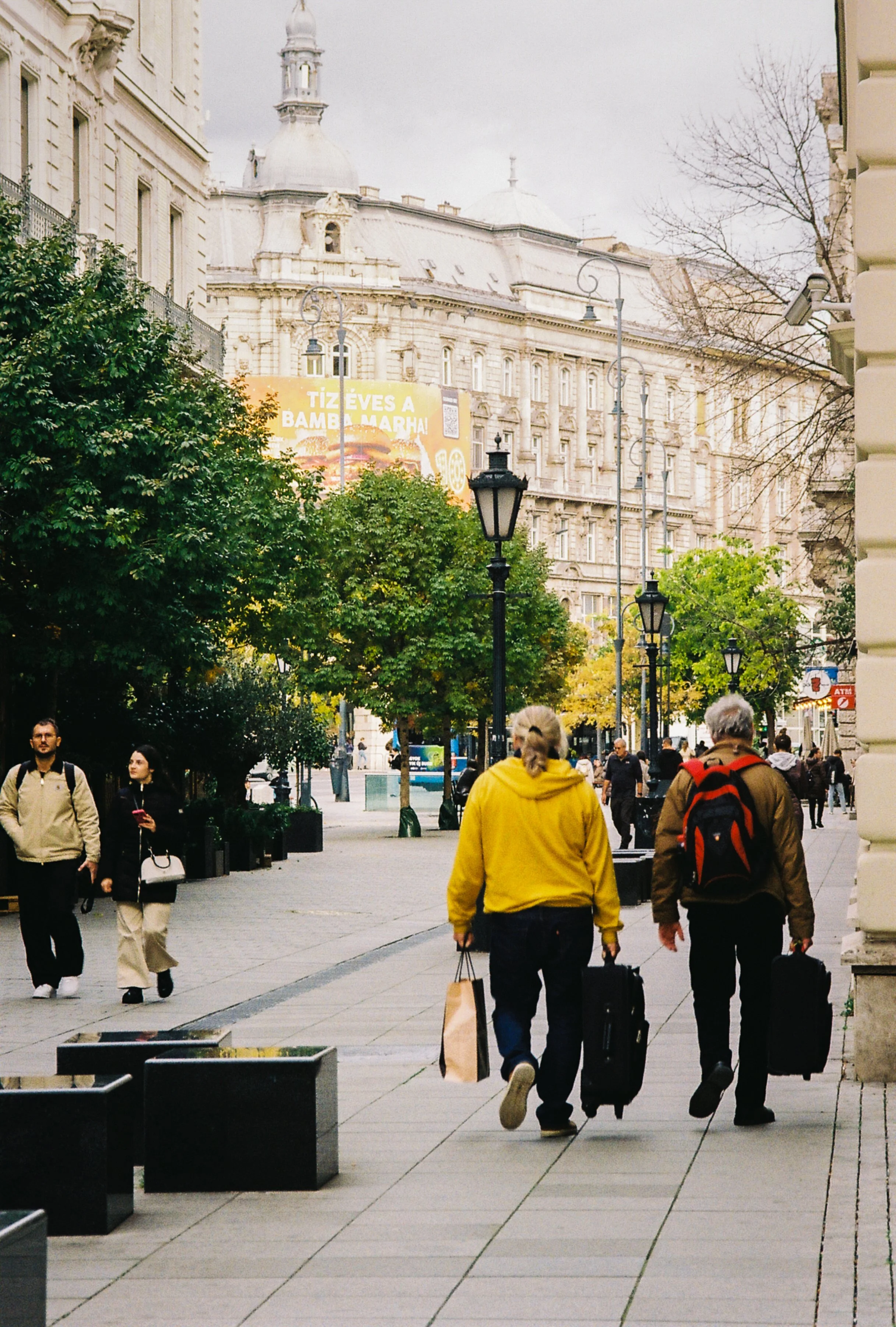 People walking on a city sidewalk among trees and street lamps with historic buildings and a billboard in the background.