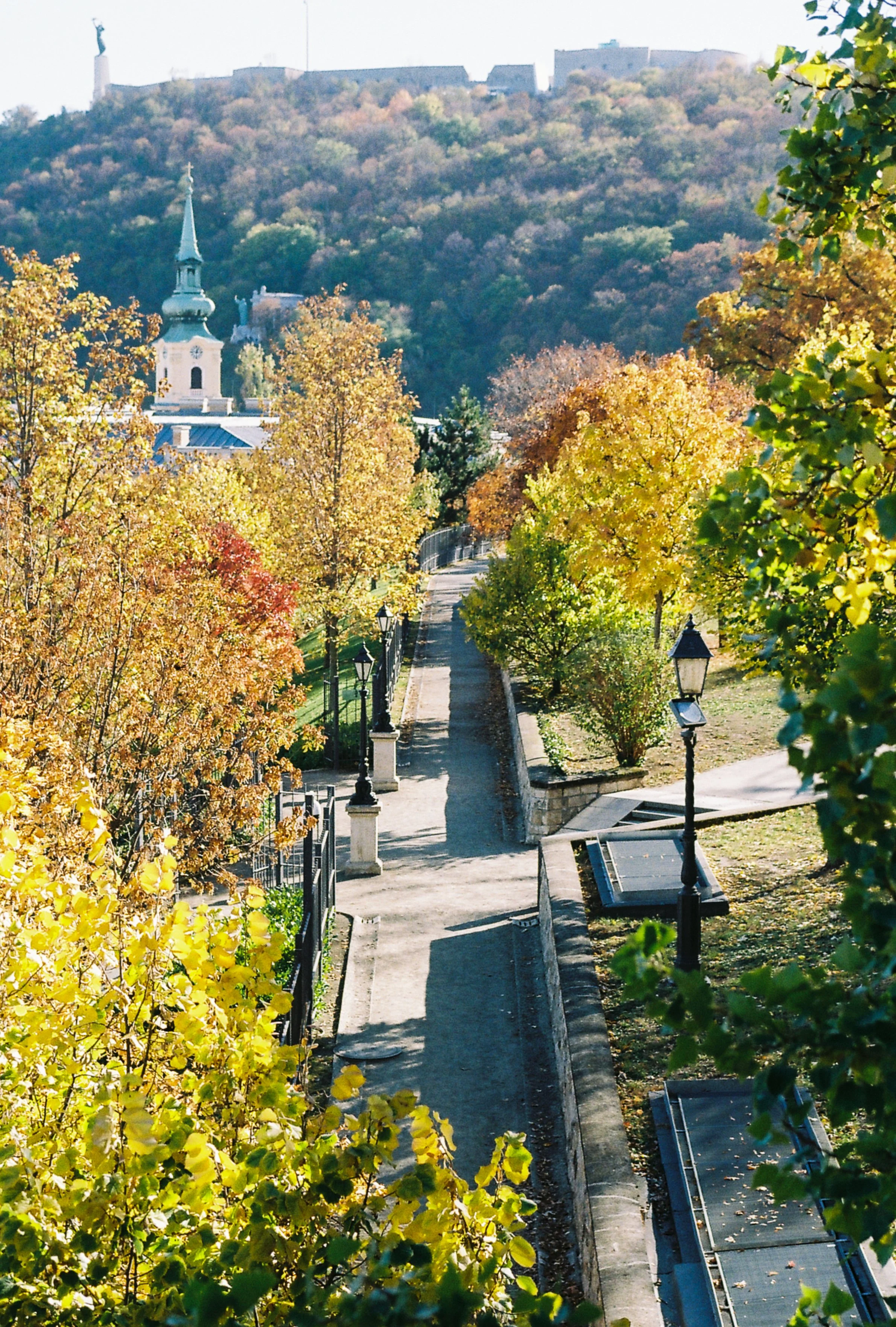 A scenic view of a pathway with autumn foliage, lampposts, and a church steeple in the distance, set against a hill with buildings on top.