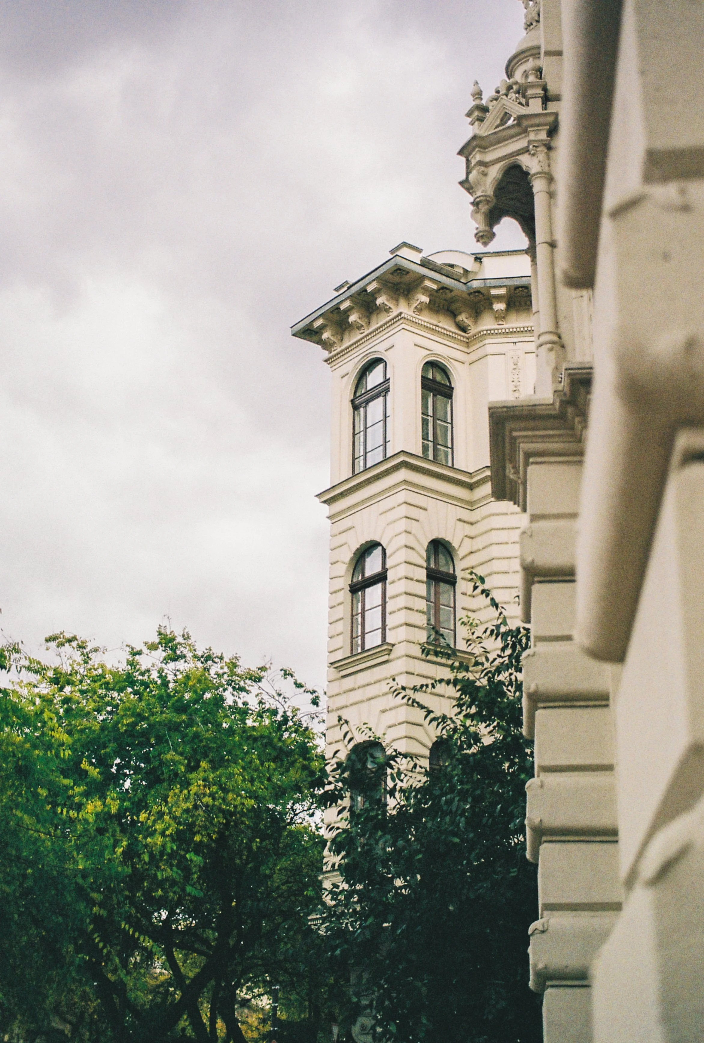 Close-up of an ornate beige historic building with tall arched windows and decorative architectural details, partially obscured by green trees, under a cloudy sky.