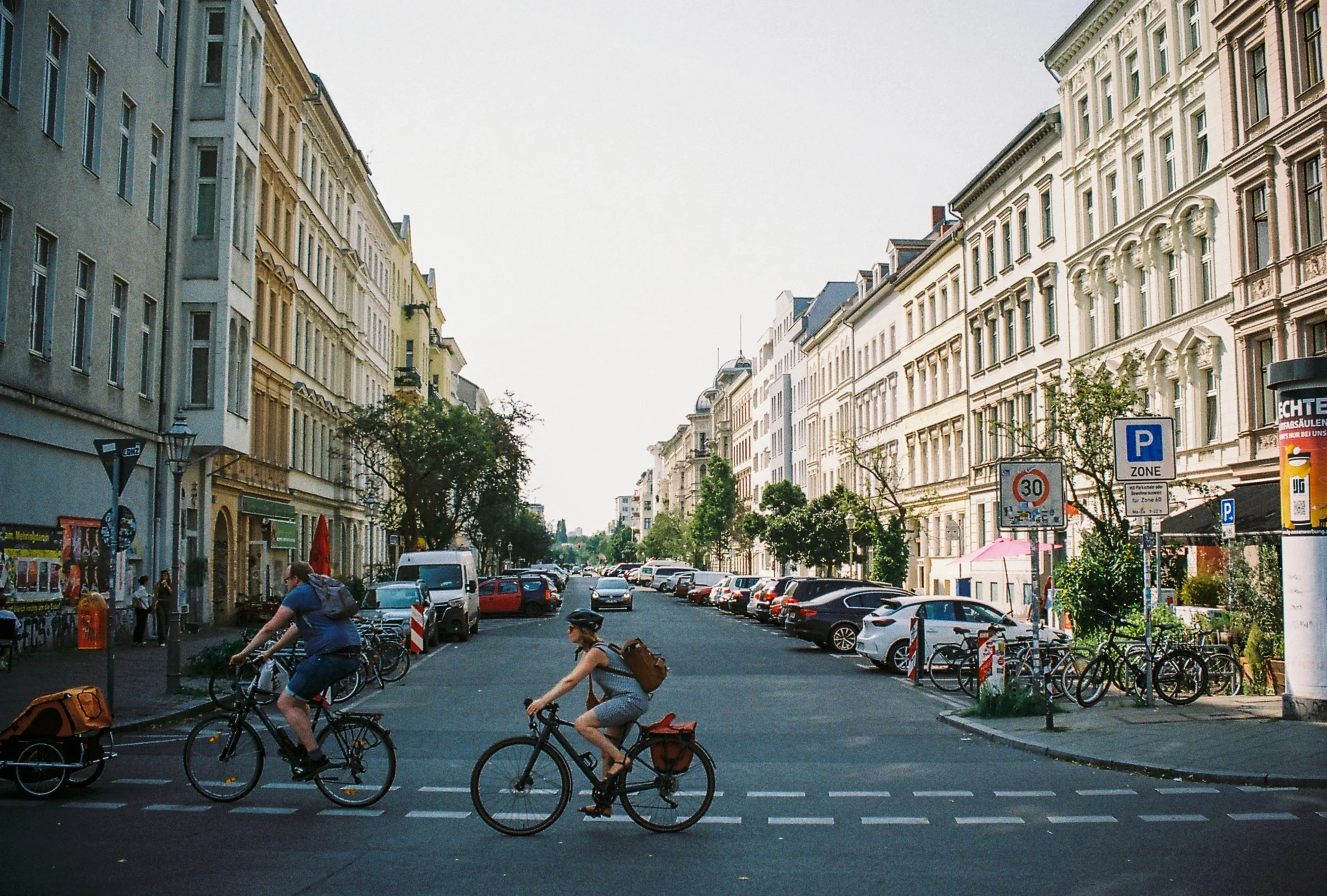 A city street with parked cars and bicycles, with buildings on both sides and a few pedestrians.