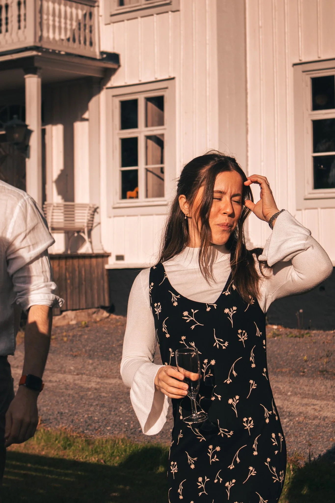 A woman appears to be in discomfort or pain, holding her head with one hand, while holding a glass of champagne in her other hand, standing outdoors in front of a light-colored house.