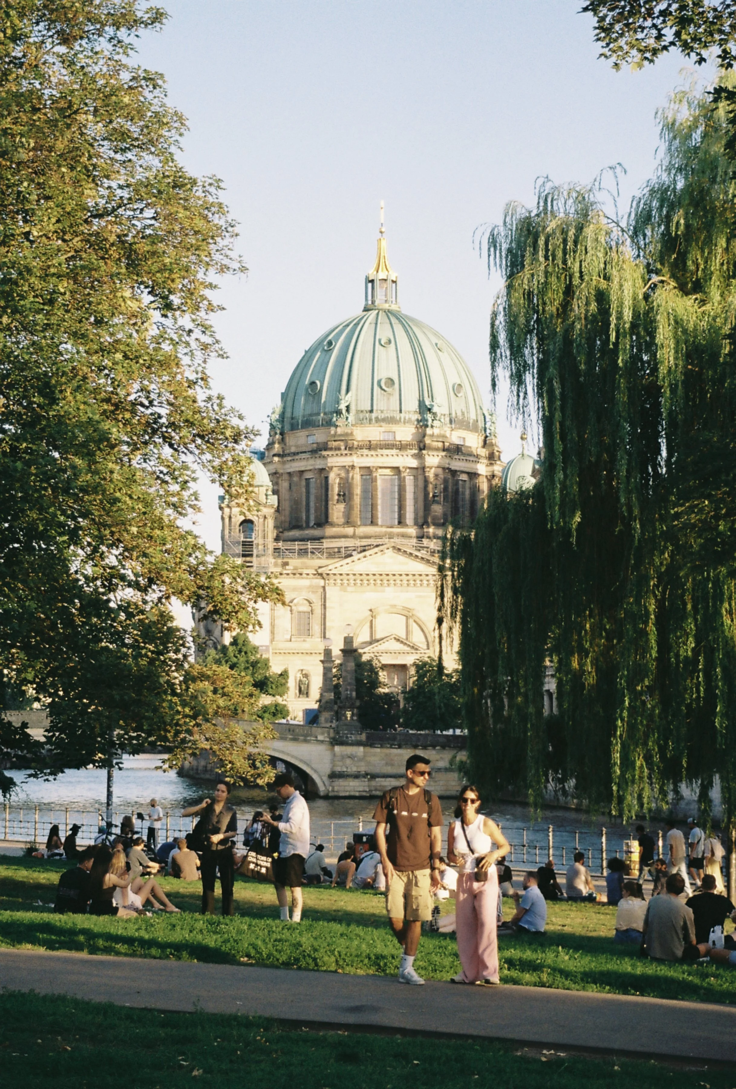 People gathered in a park with a historic domed building in the background, possibly a cathedral, along a riverbank with trees lining the area.