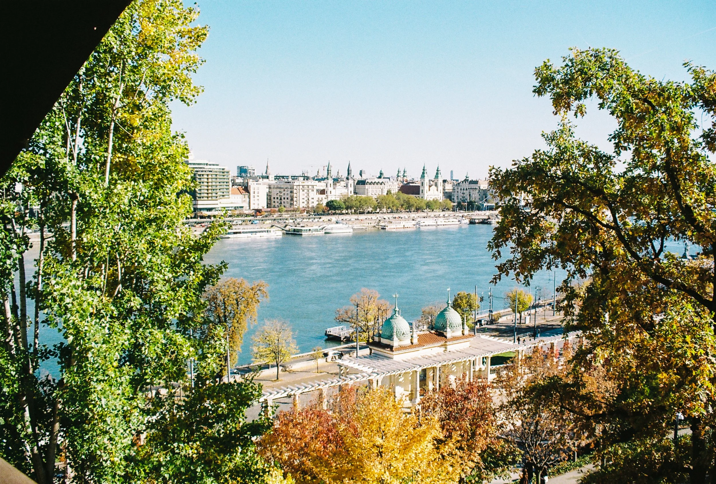 View of a river with boats along the shoreline, surrounded by trees with autumn leaves, and a city skyline with historic buildings and spires in the background.