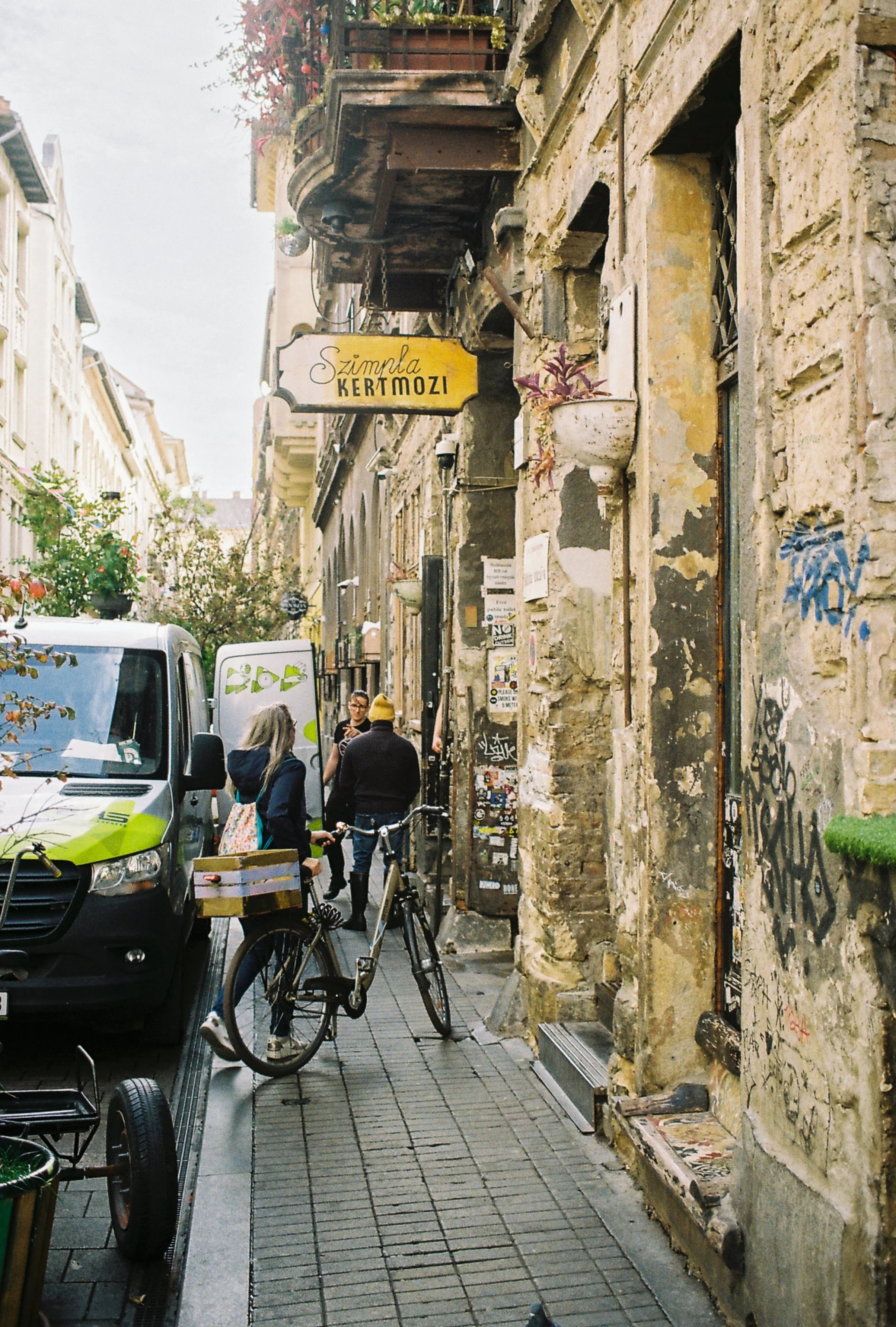 A narrow street in an old city with a weathered building facades, graffiti, and a few people, including two women and a man, standing near a bike and a van, with a yellow sign reading 'Szimple Kertmozi' hanging above.