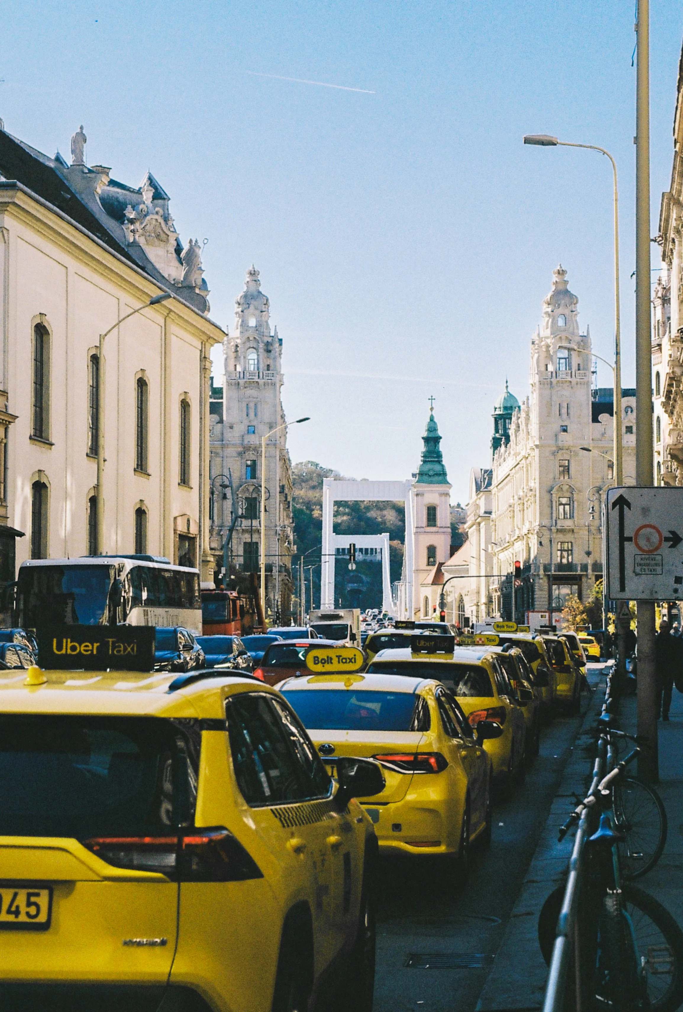 City street scene with yellow taxis, Uber taxis, and a line of cars parked on the side. In the background, tall ornate buildings and a hill with a church and a decorative archway are visible.
