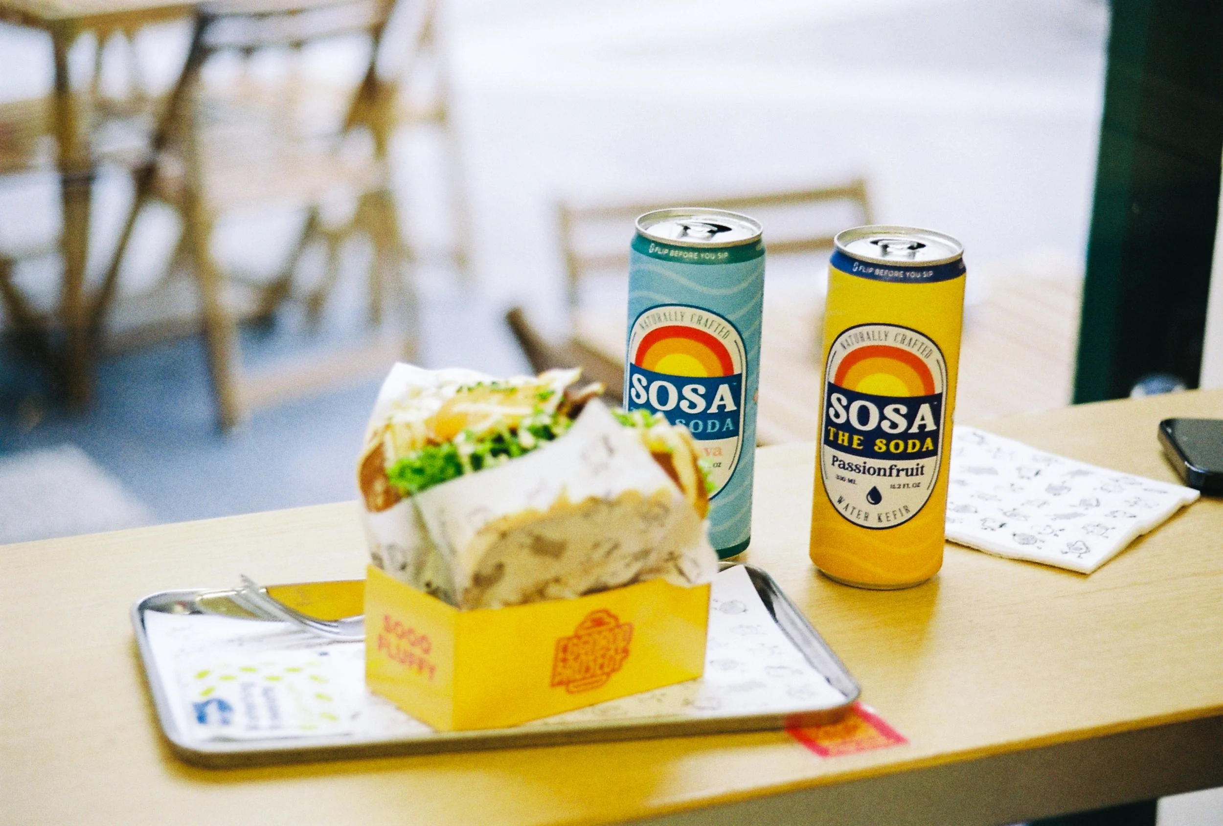 A tray with a sandwich and two cans of soda placed on a wooden table, with a background of wooden furniture and a bright, outdoor setting.