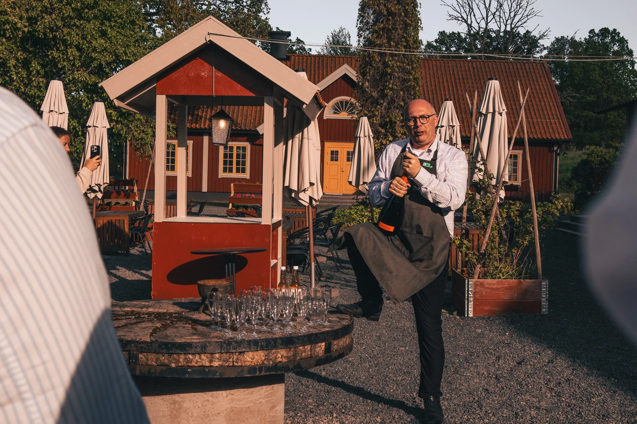 A man in a white checkered shirt and black apron standing outdoors next to a round stone table with glasses and bottles, holding a champagne bottle in one hand and a corkscrew in the other, with a rustic red house with yellow doors and surrounded by 