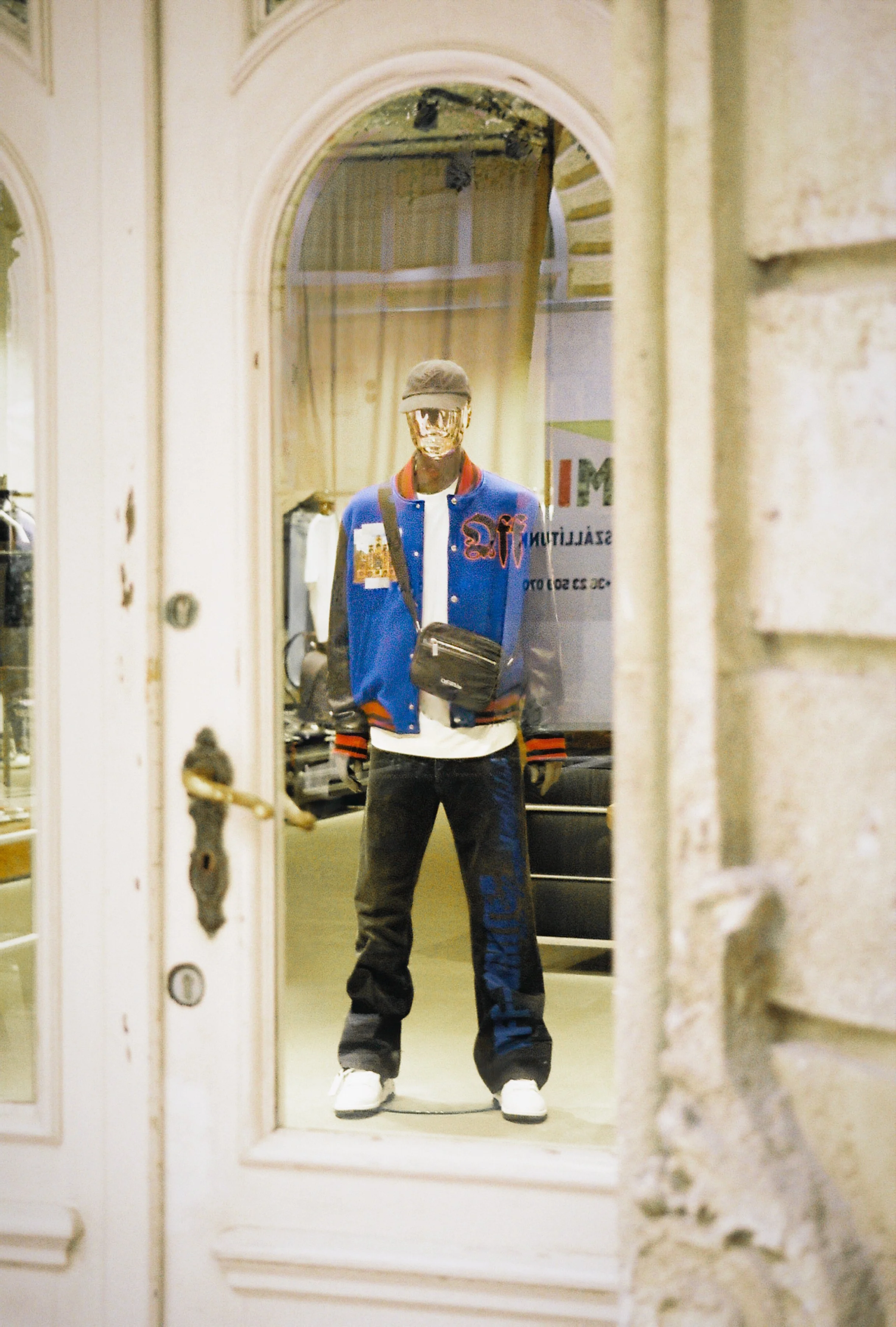 A mannequin is seen through a door mirror, dressed in a blue and red varsity jacket, black pants with blue lettering, white sneakers, a black cap, and a black shoulder bag.