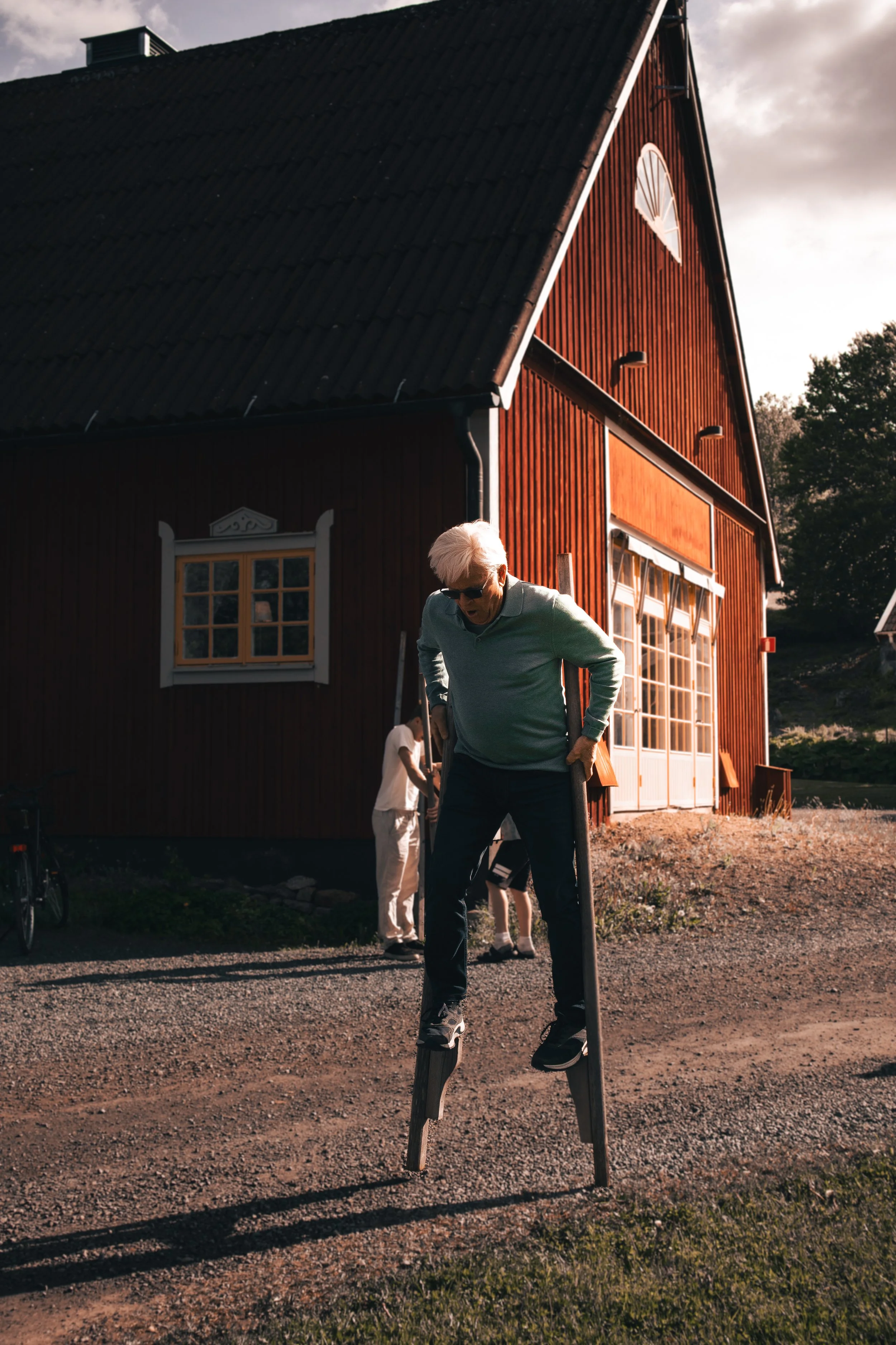 A man on stilts walking on a dirt path outside a red barn during sunset.