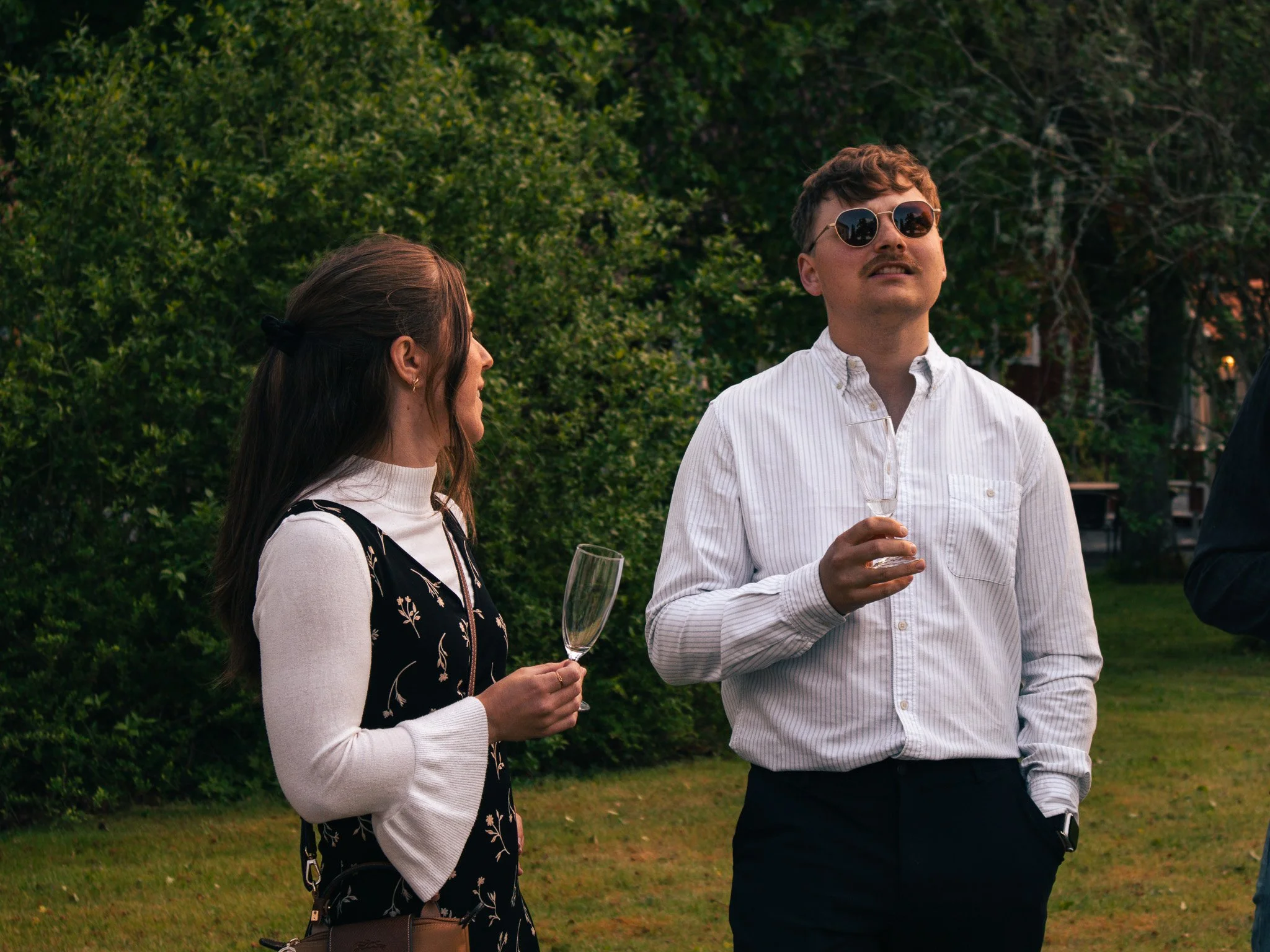 A man wearing sunglasses and a white dress shirt holding a glass of wine, standing outdoors with a woman who has long brown hair, wearing a patterned black dress with white sleeves, holding a glass of wine, at a social gathering in a park or garden s