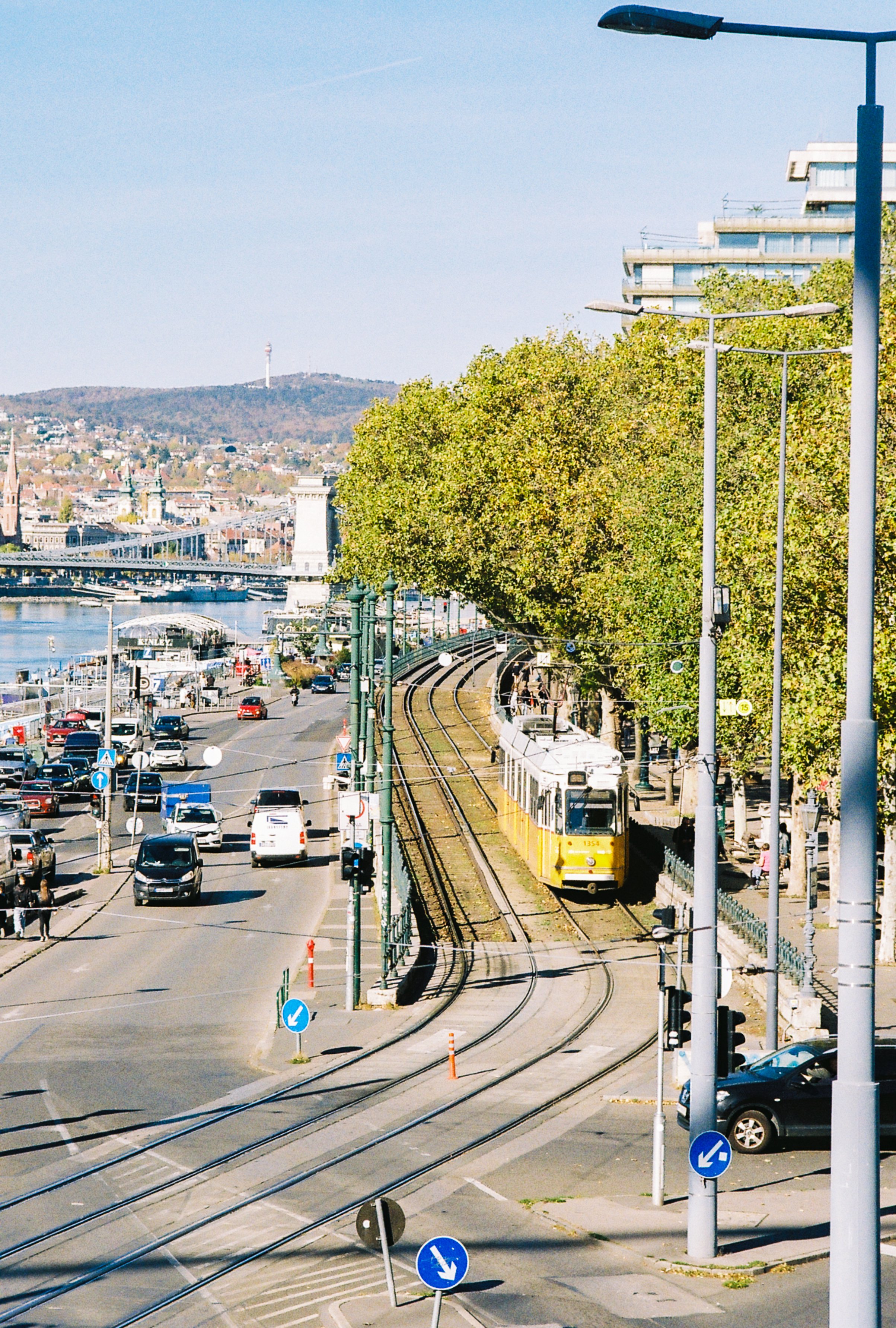 A city street with a yellow tram, cars, and trees alongside the tracks. The river and a bridge are visible in the background, with hills and buildings beyond.