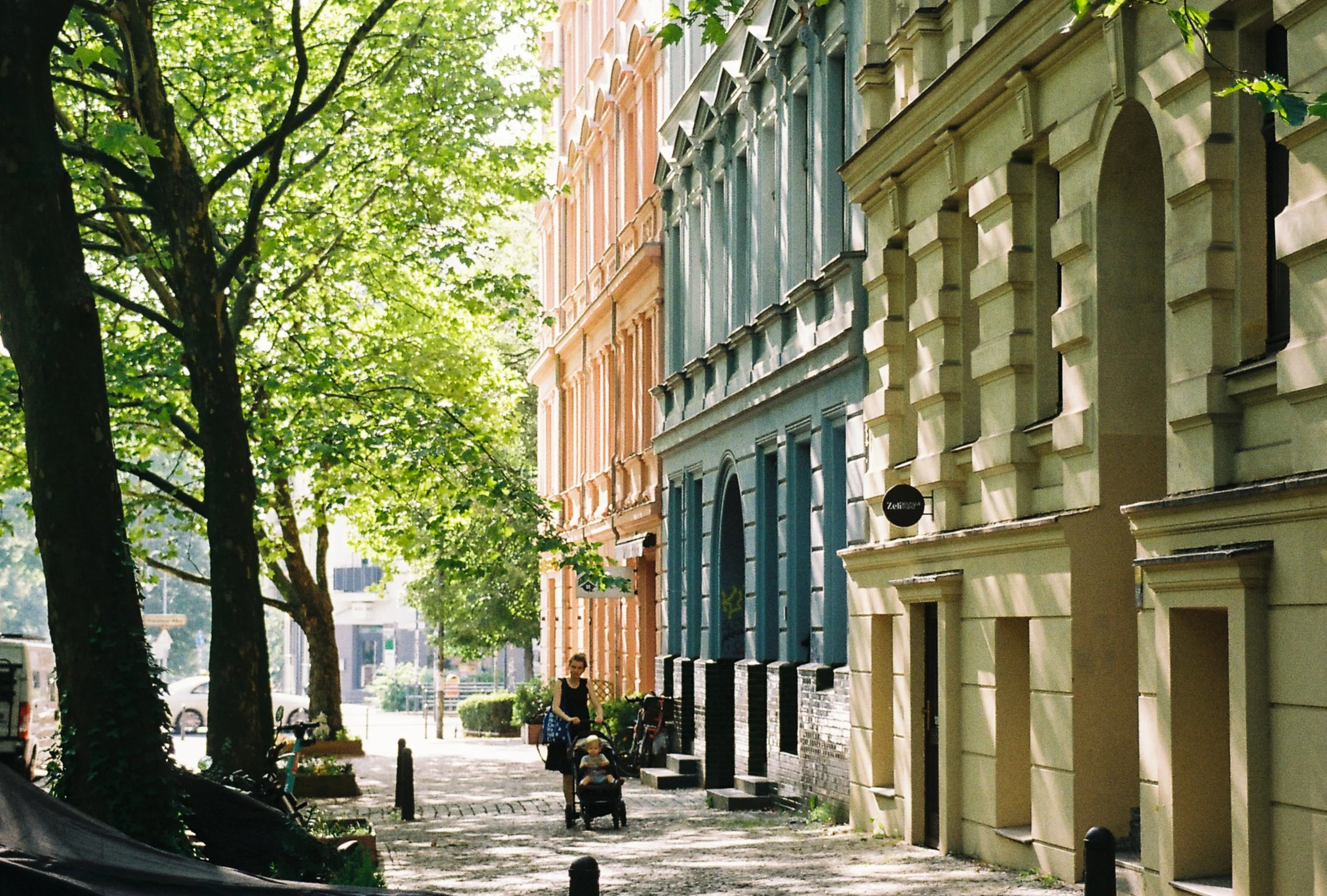 A person walking with a stroller along a shaded sidewalk lined with colorful buildings and large trees.