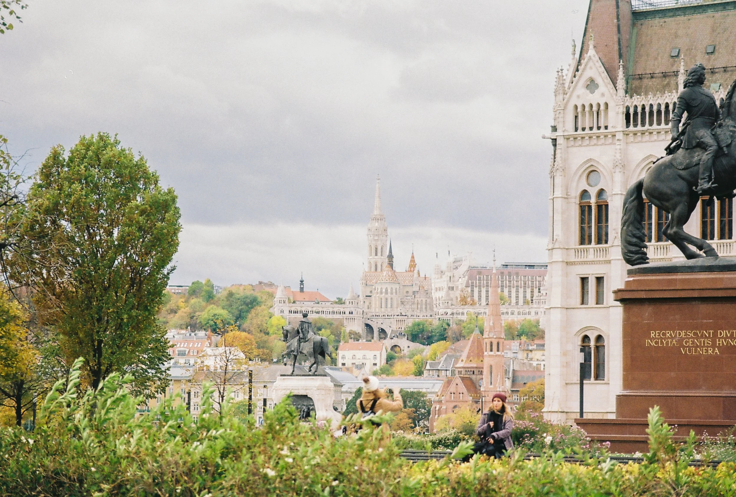 Cityscape view of Budapest, Hungary with historical buildings, statues, and trees, under cloudy sky.