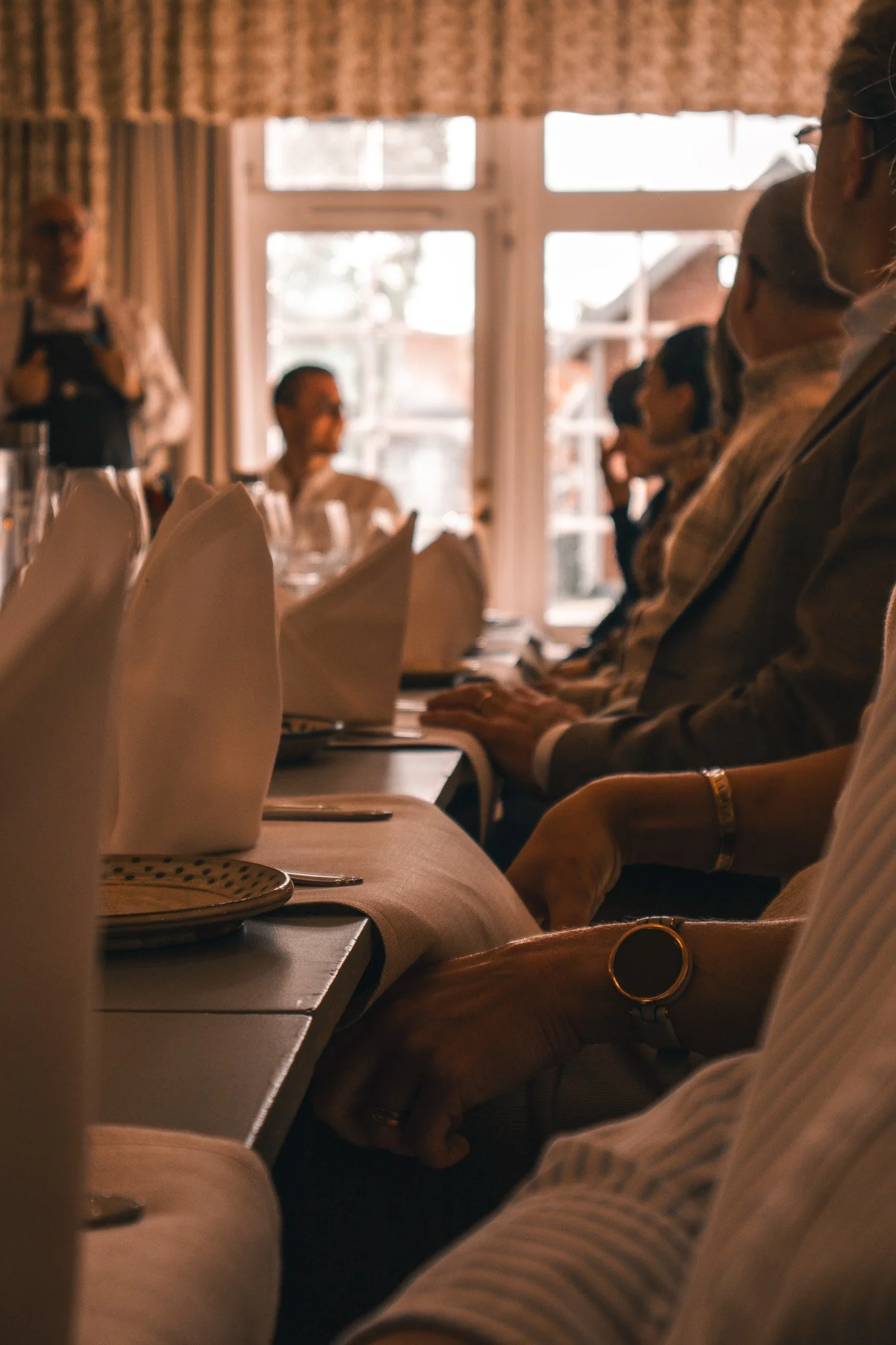 People seated at a dining table in a restaurant, with folded napkins, plates, and cutlery, during daytime, some individuals wearing face masks, with a waiter standing in the background near large windows.