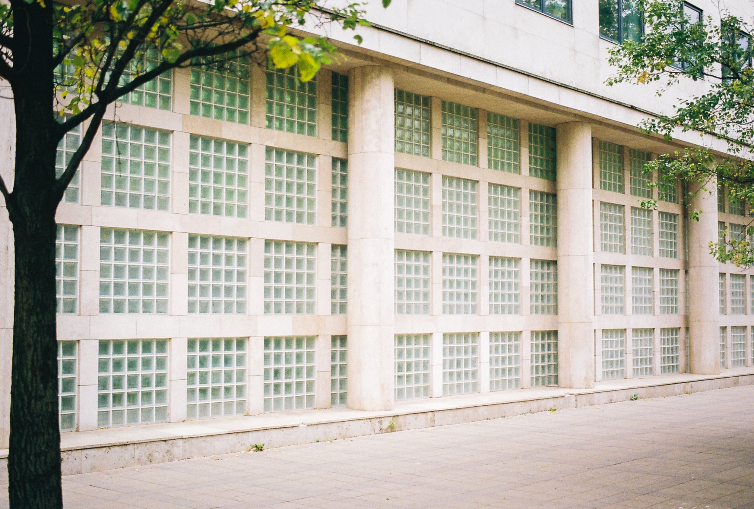 A modern building with large glass block windows, beige stone pillars, and a sidewalk in front, with green trees along the street.