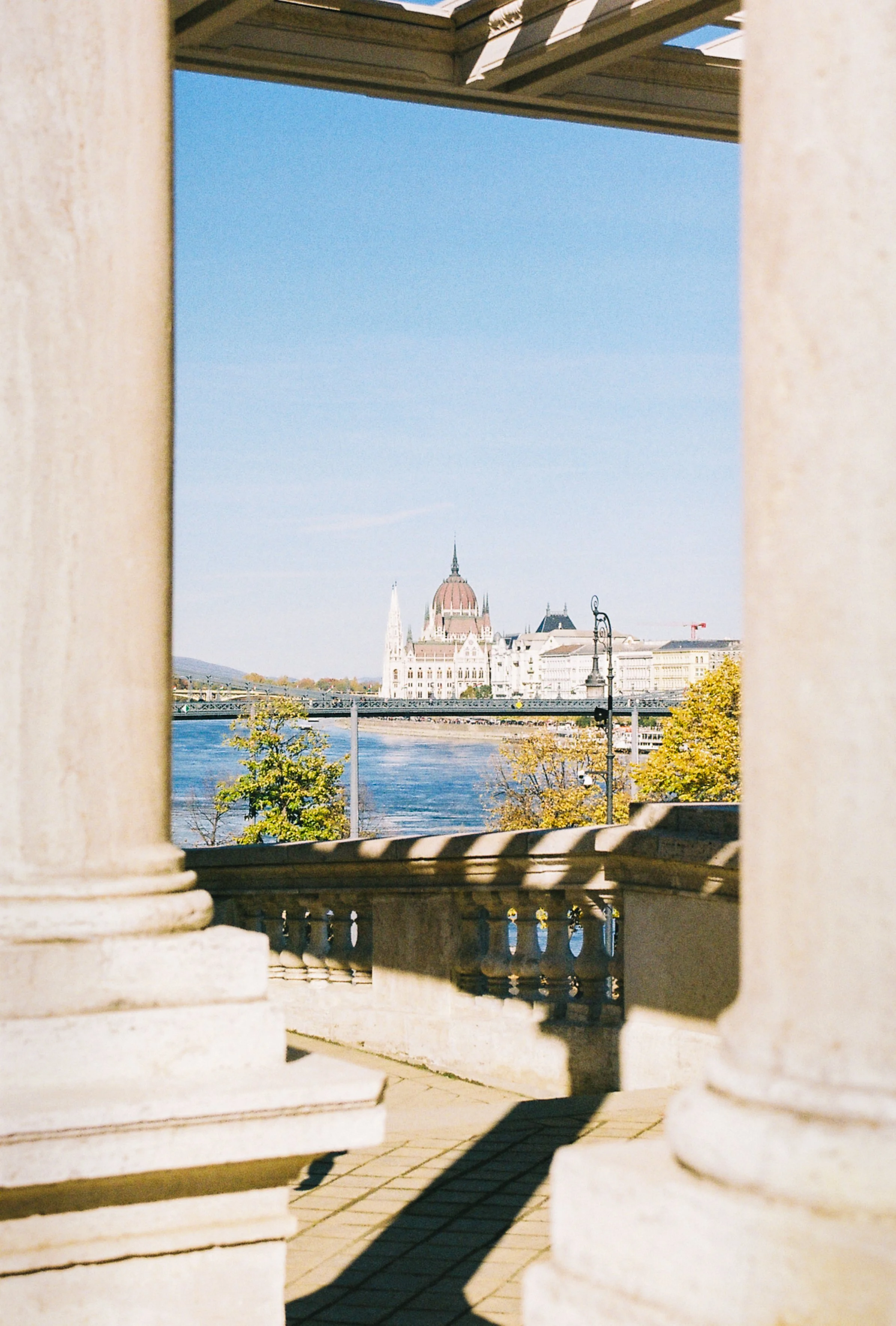 View of the Hungarian Parliament Building across the Danube River, framed by stone columns and a balcony in Budapest, Hungary.
