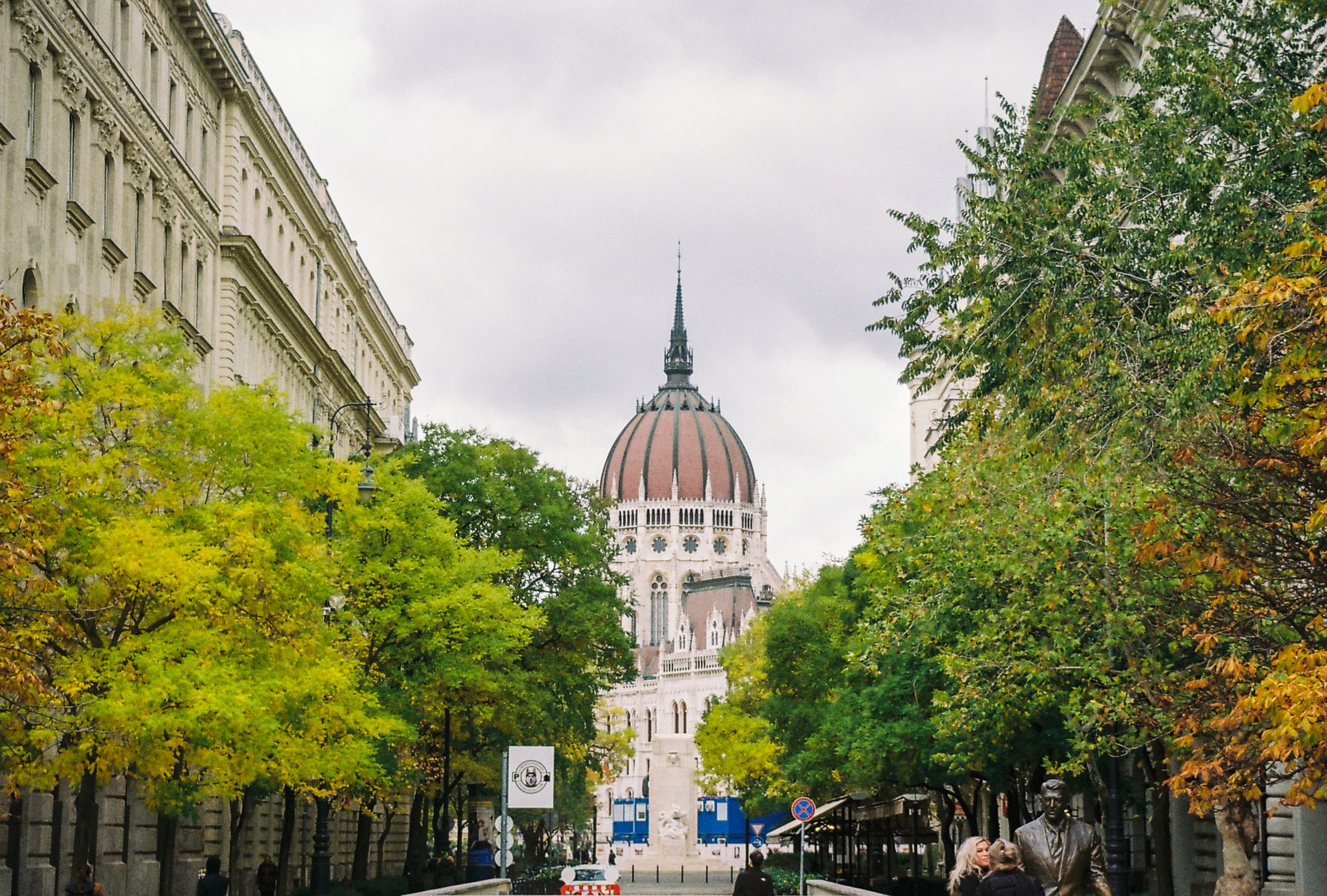 A city street scene with green and yellow trees lining the sidewalk, a historic building with a domed tower in the background, and pedestrians walking along the street.