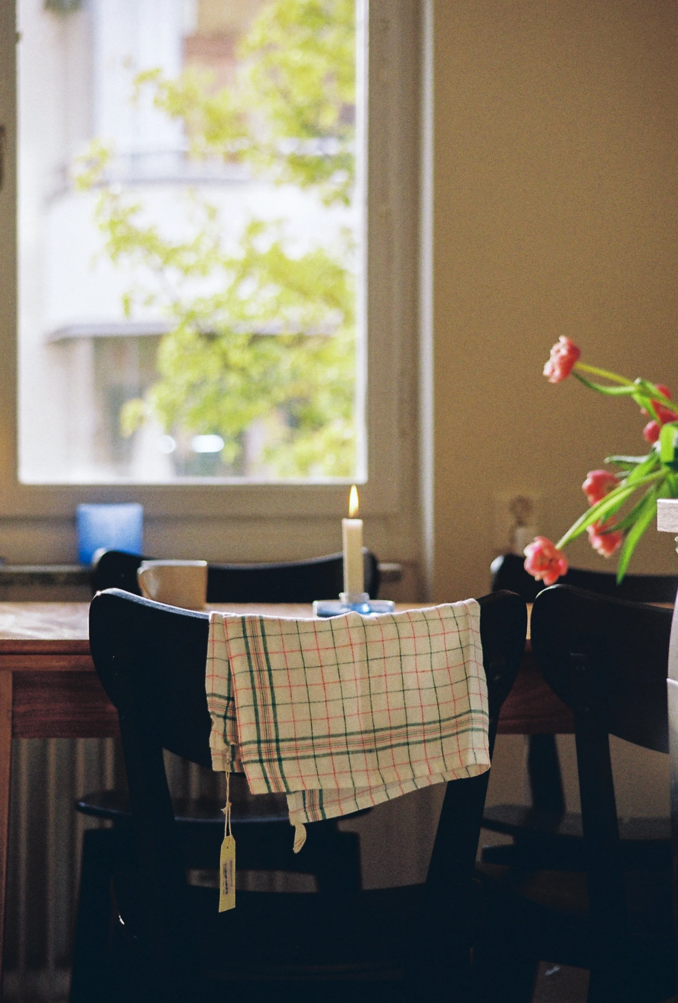 A dining room scene with a wooden table and four black chairs; one chair has a checkered towel draped over the backrest. A lit candle is on the table. There is a window in the background showing greenery outside, and a vase with pink flowers on a sid