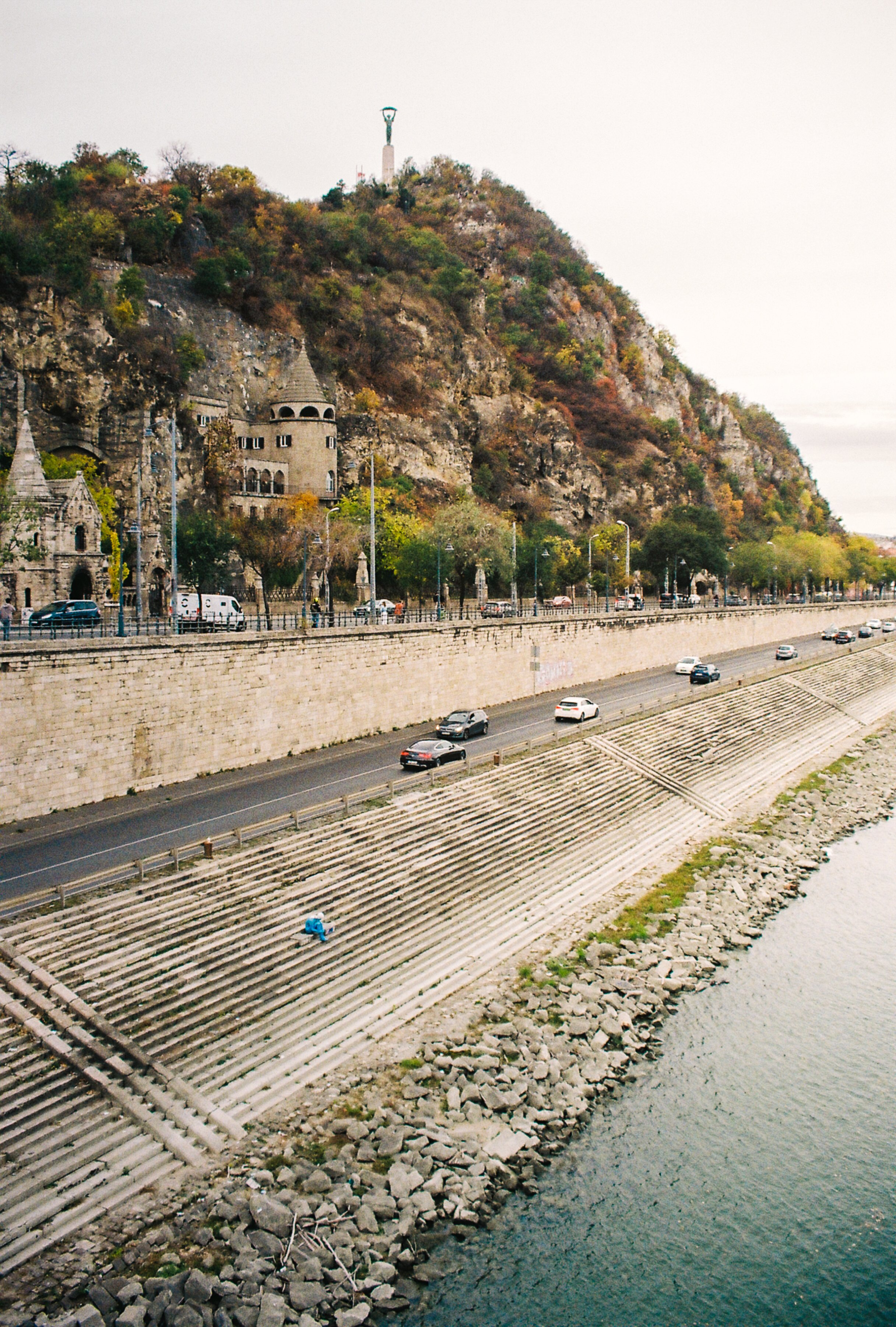 A riverside scene with a steep embankment, stone steps, and a person sitting by the water, with a mountain and a statue in the background.