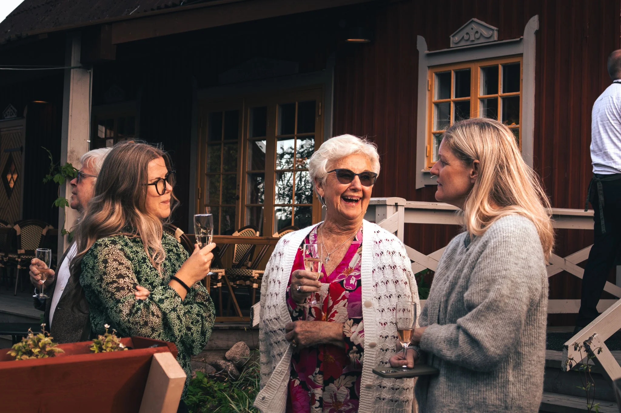 Four women and one man at an outdoor gathering, holding champagne glasses, engaged in conversation, with a rustic wooden building in the background.