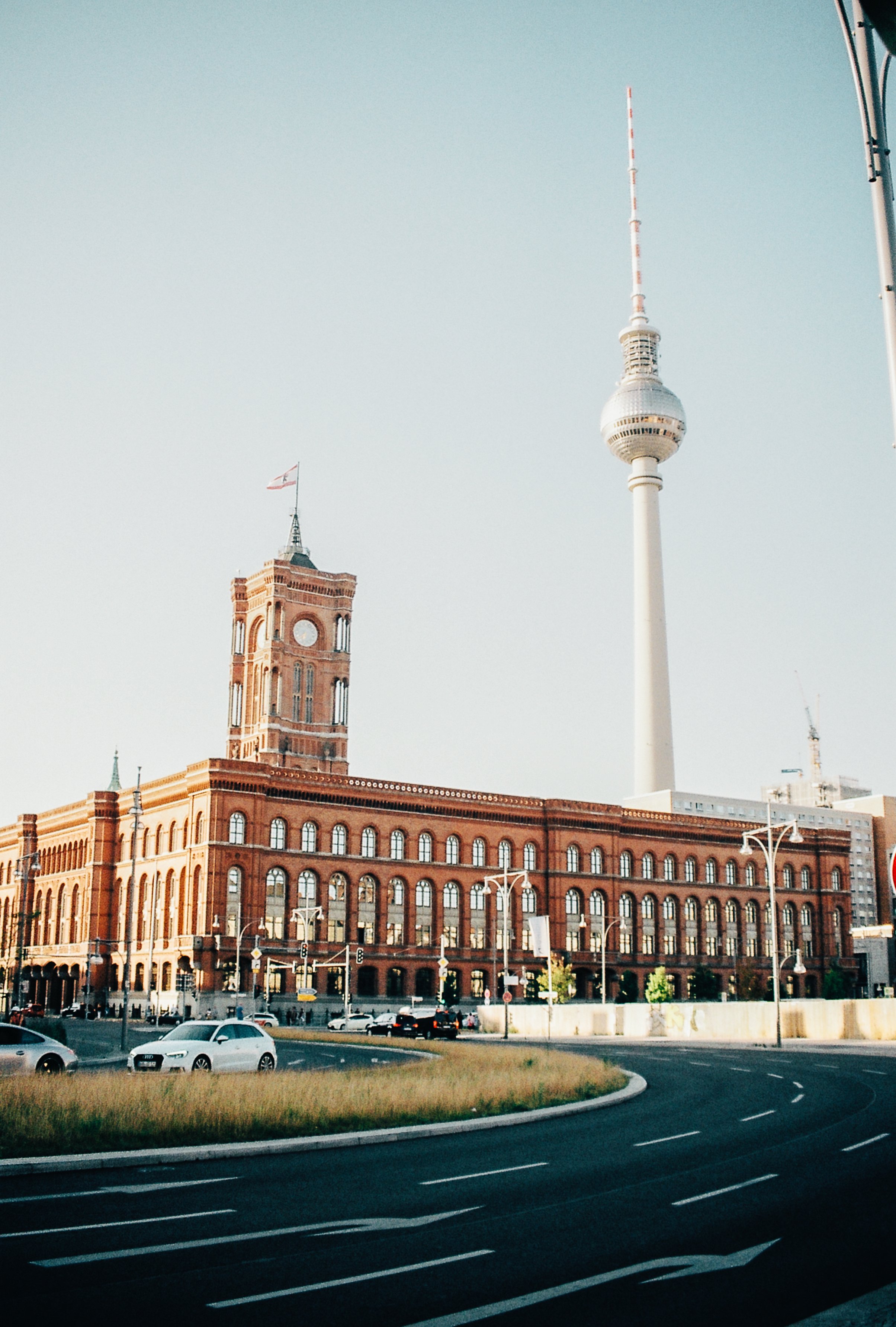 View of the Berlin TV Tower and the red brick Rotes Rathaus (City Hall) in Berlin, Germany, with cars on a curved road in the foreground and a clear blue sky above.