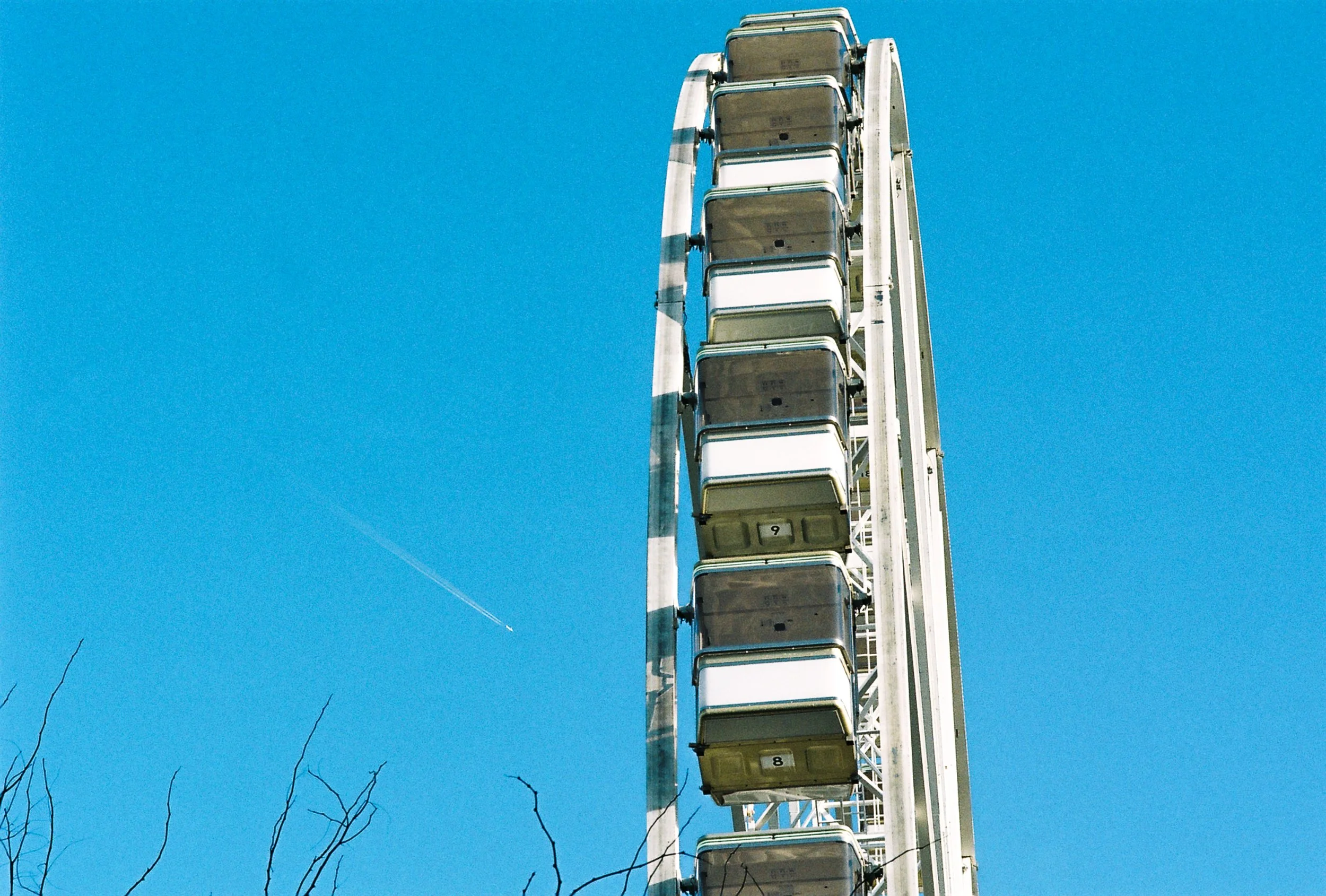 A close-up view of a Ferris wheel with numbered gondolas against a clear blue sky, with bare tree branches at the bottom of the image.