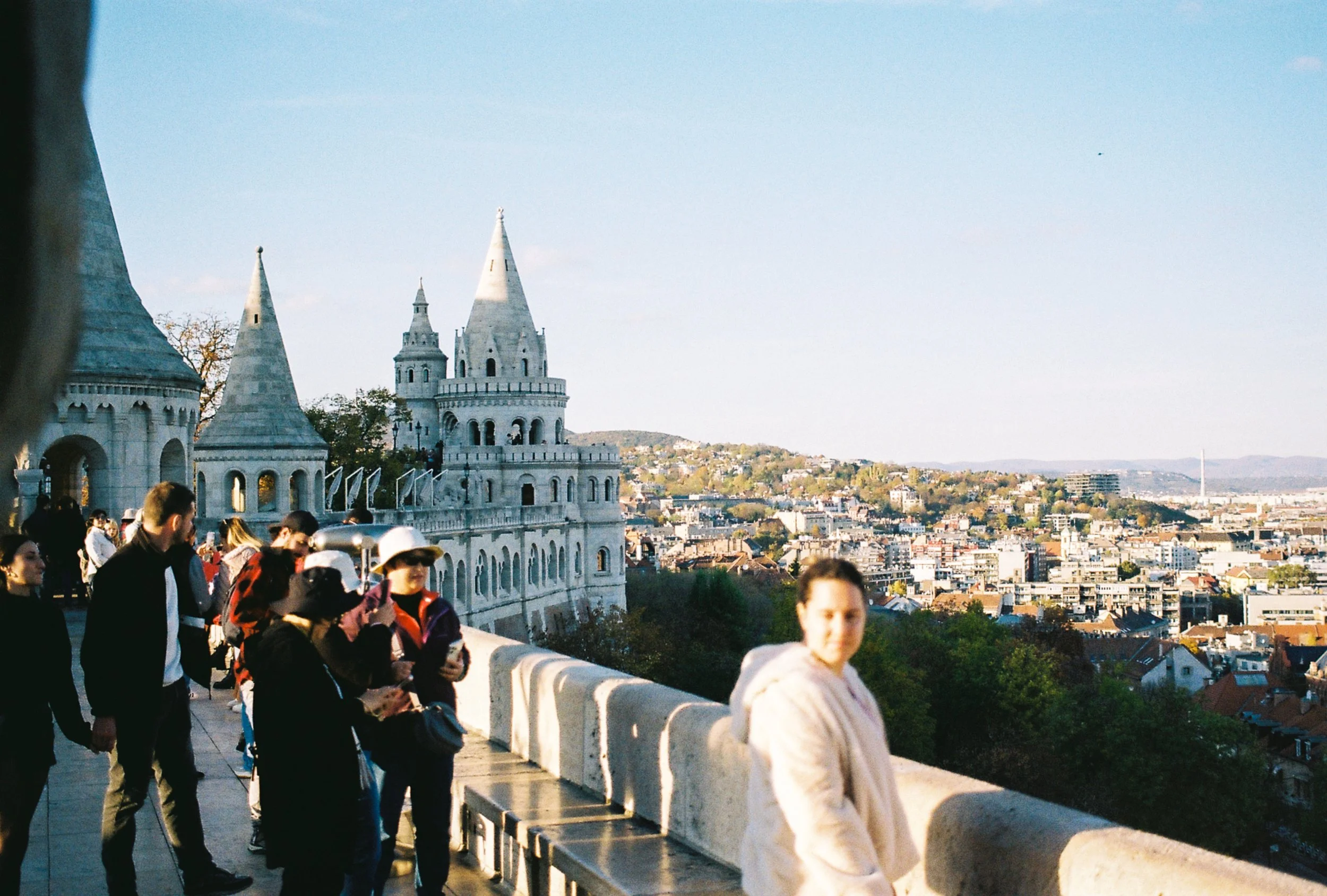 Tourists and visitors standing on a ledge overlooking the city of Budapest, Hungary, with a view of the Fisherman's Bastion, a neo-Gothic and neo-Romanesque fortress with turrets, in the background during daytime.