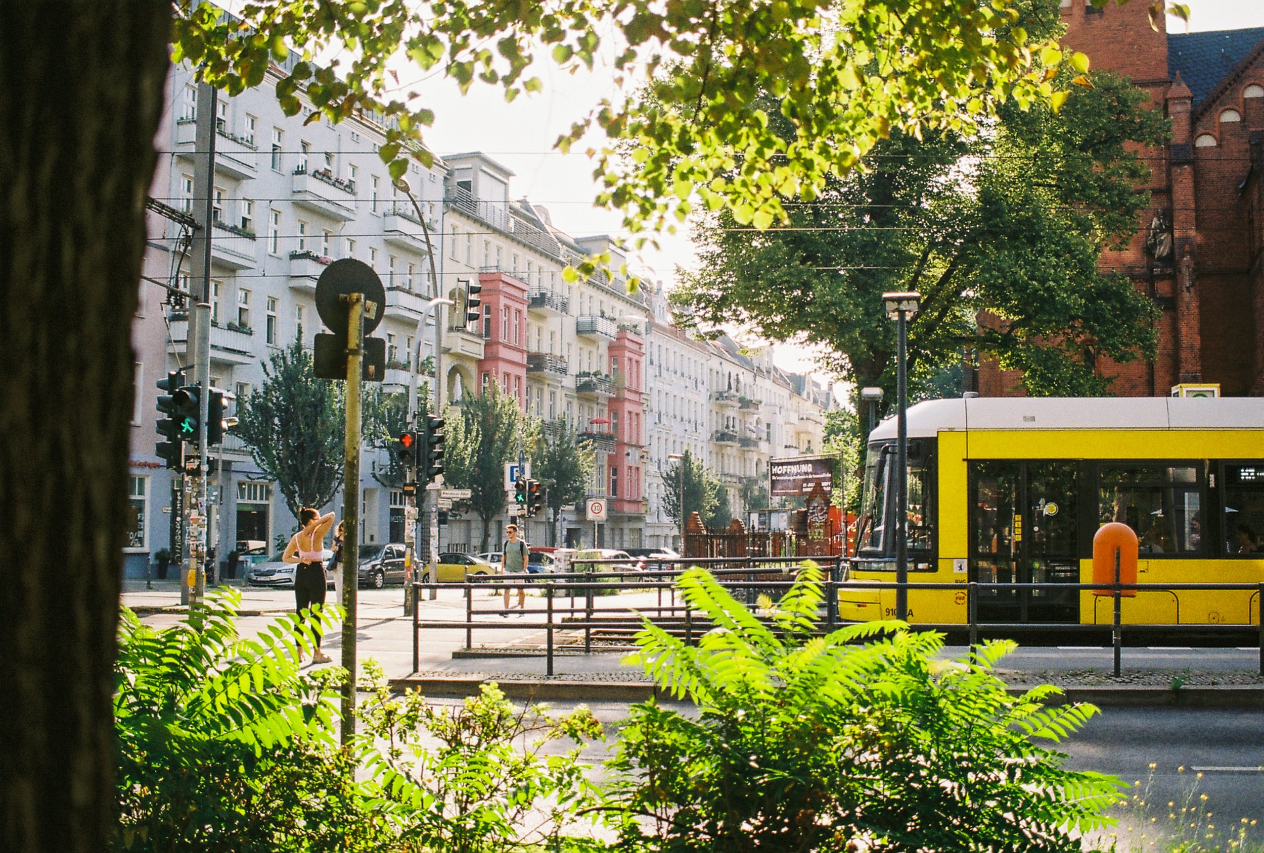 A street scene in an urban area with a yellow tram, pedestrian crossings, and buildings with balconies. Green graffiti-covered trash bins are visible, and a woman is standing at the crosswalk as another person walks by. Trees frame the scene, and sun